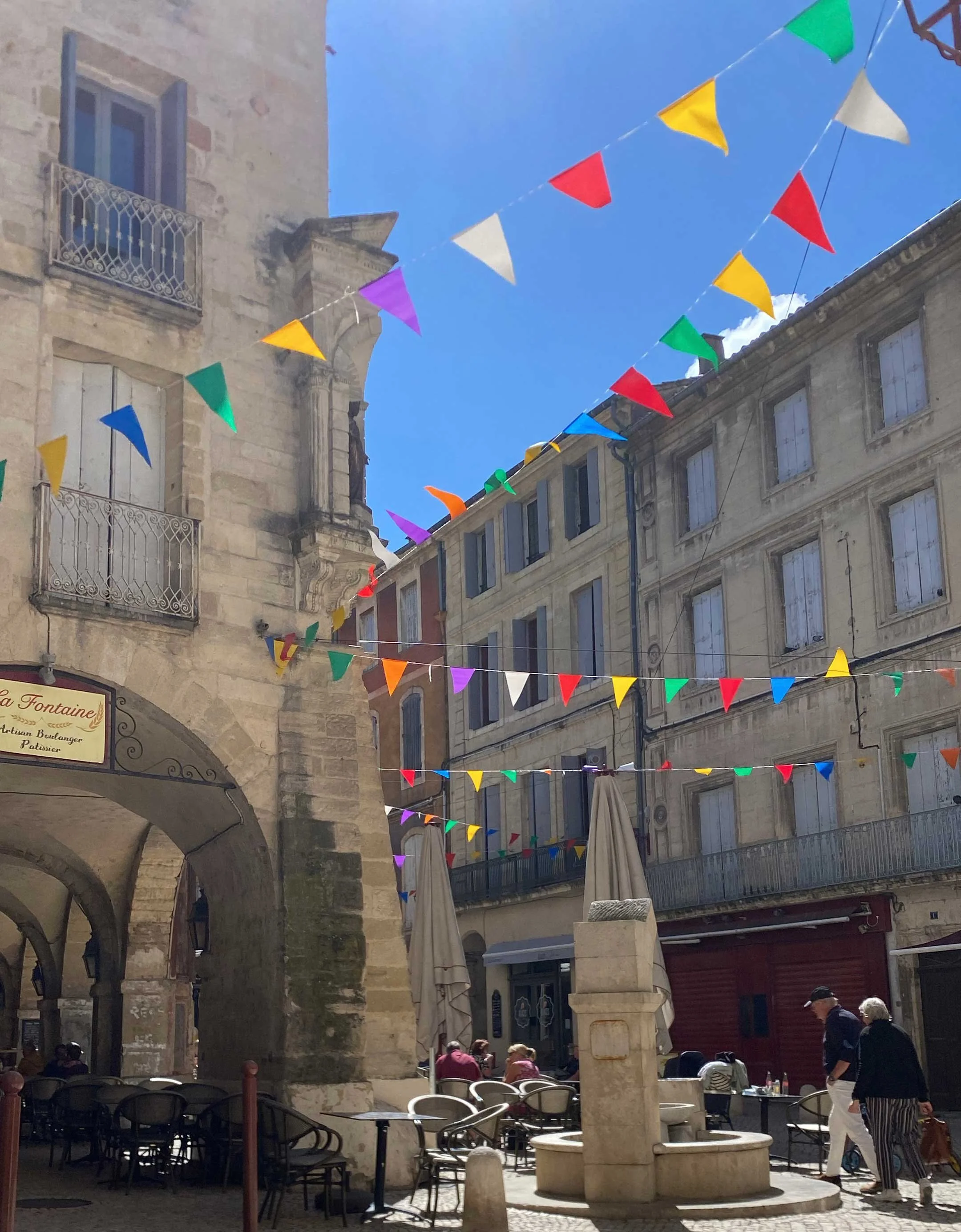 Colorful bunting flags hanging across a European city square under a blue sky, with outdoor cafe tables and people walking.