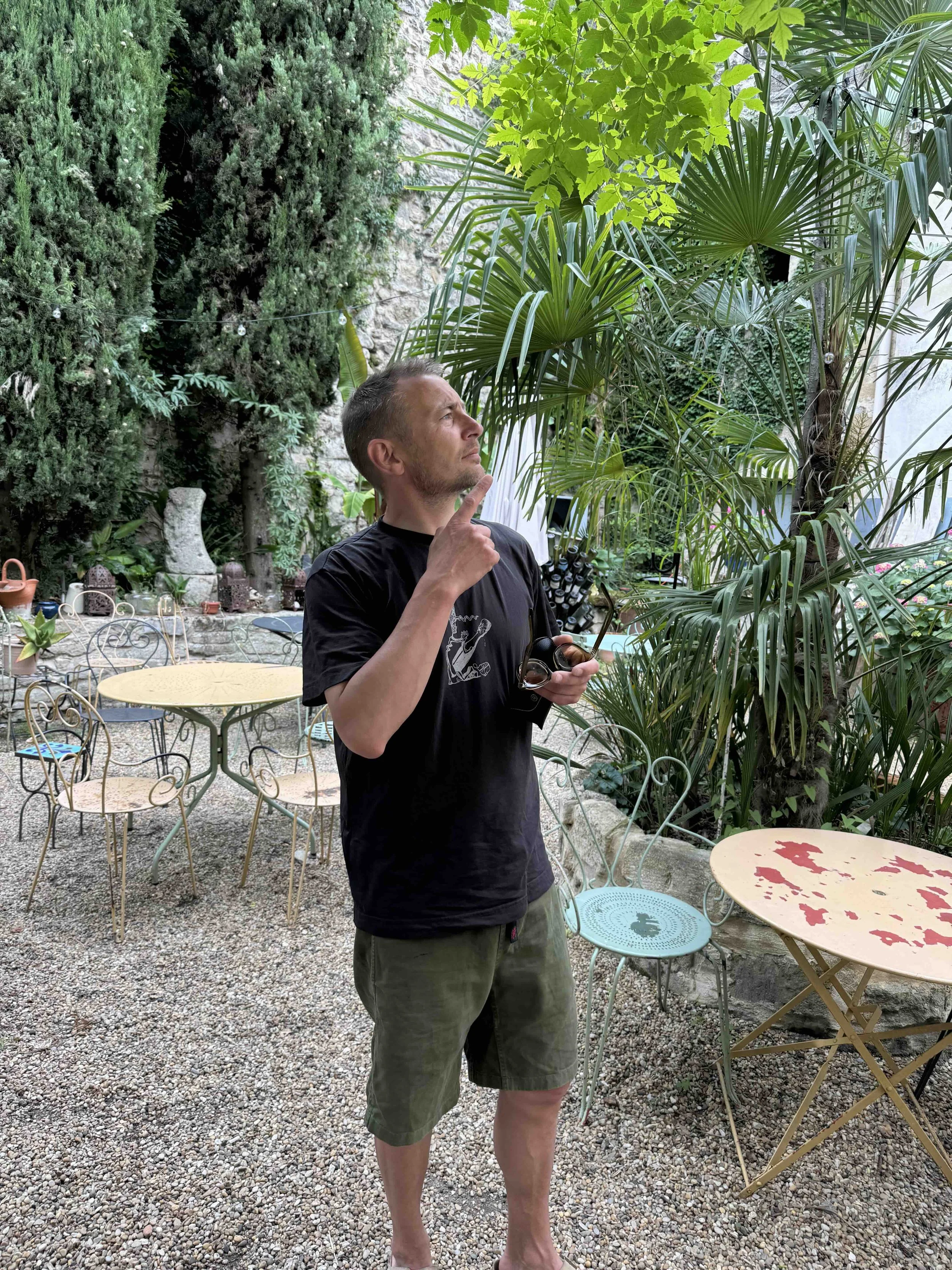 Mads lønnerup standing outdoors in a garden with lush green plants, including palm trees and tall shrubs, holding a camera and touching his chin, looking thoughtfully into the distance.