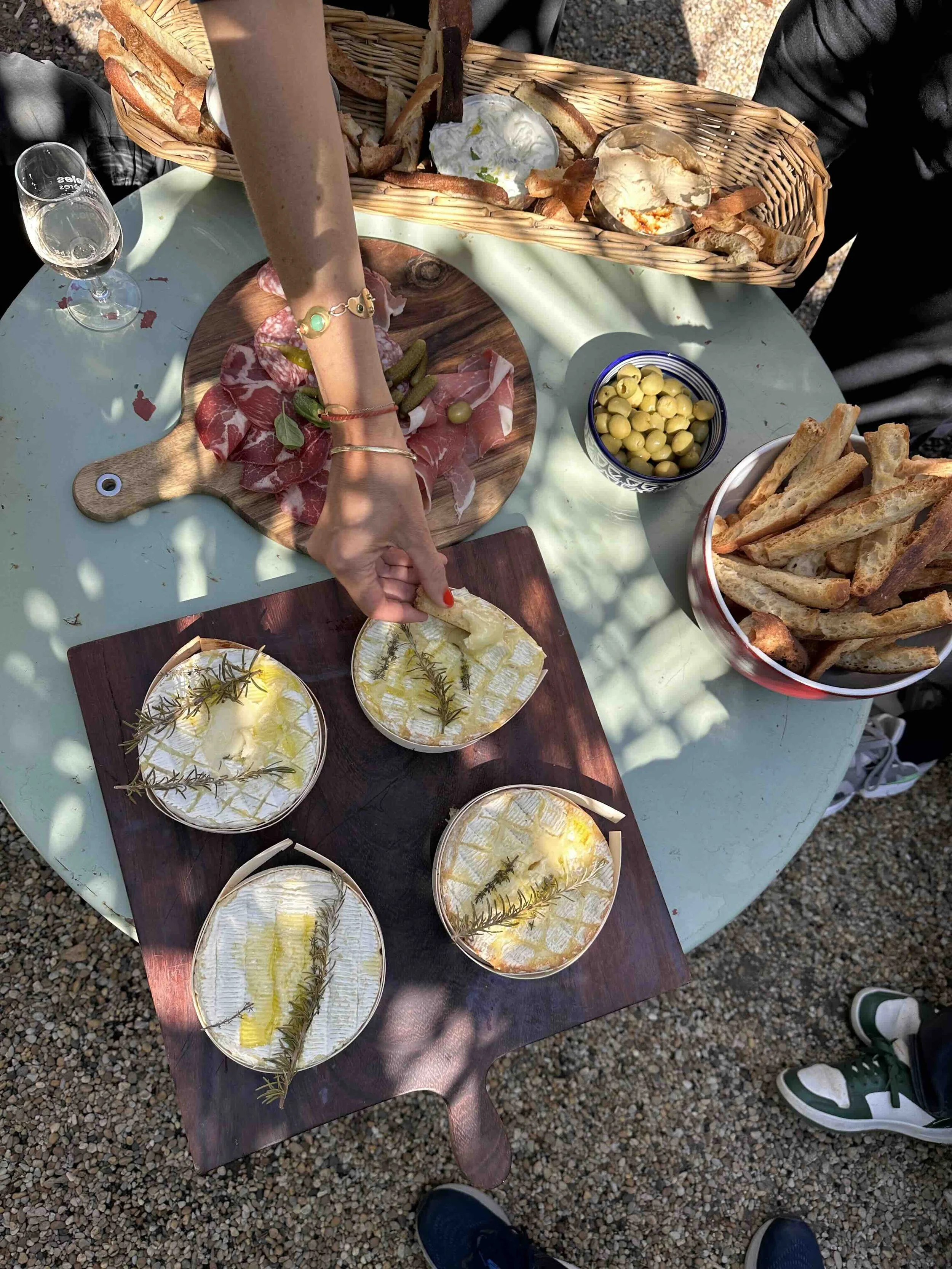 A table set with bowls of cured meats, bread, cheese, olives, and a small glass of white wine. Someone is reaching for cheese and sprigs of rosemary, with various snacks and a basket of bread on the table.