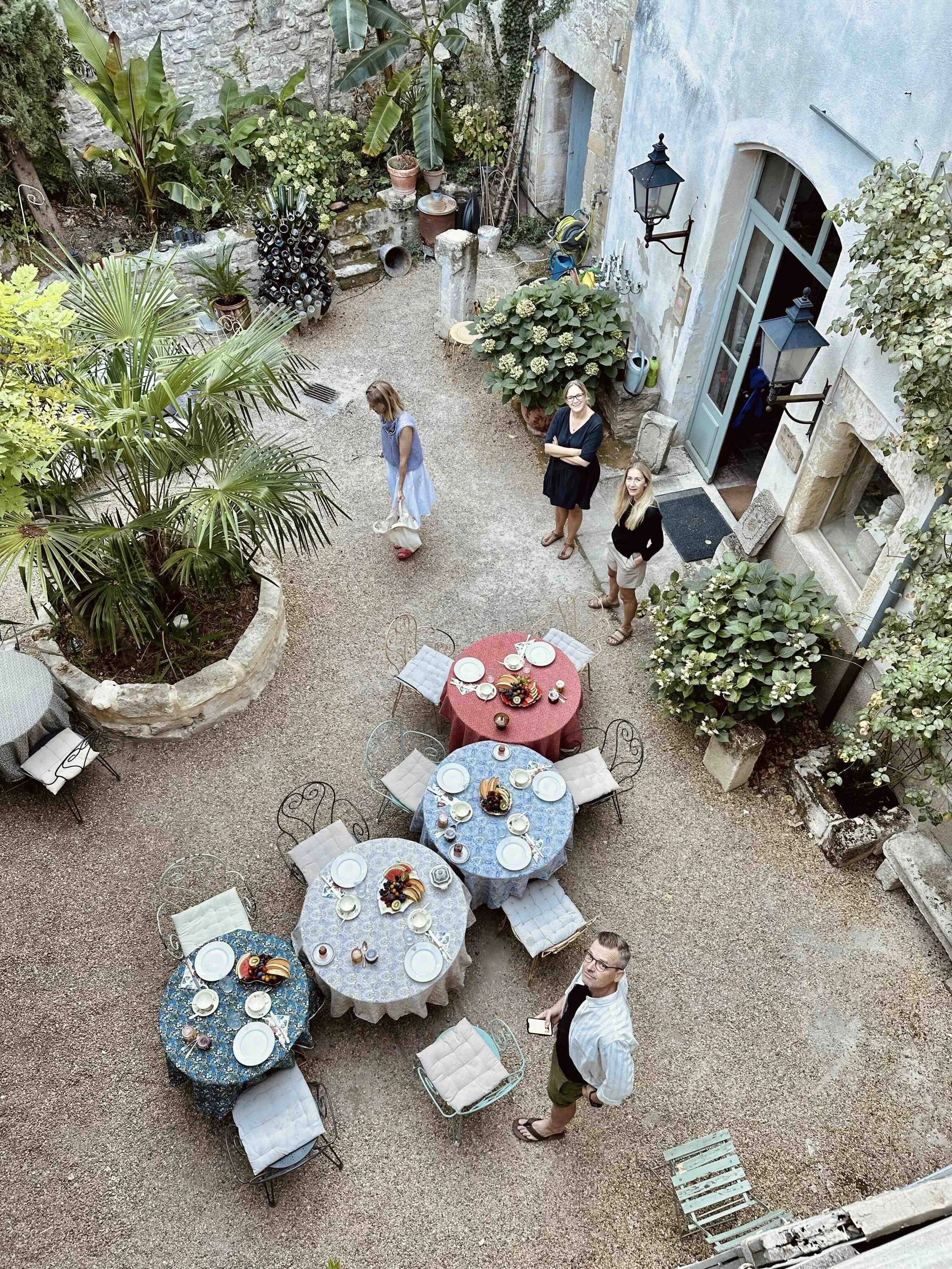 An outdoor terrace with trees and potted plants, set with several round tables covered with patterned tablecloths, plates, and food. Four people are present; one woman is standing near a door, two women are standing together, and a man is standing near a table, looking up at the camera.