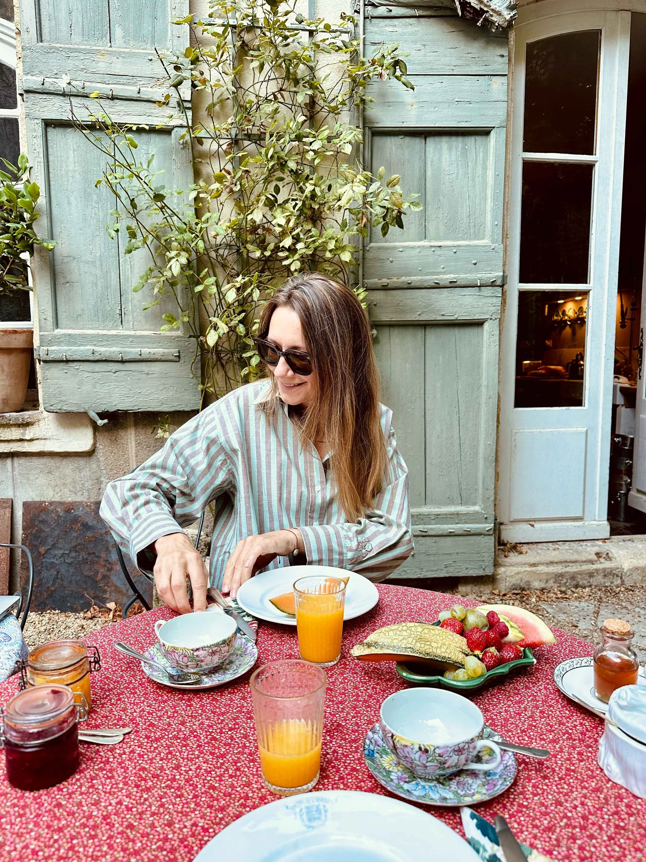 Anne Mette enjoying breakfast in the garden wearing sunglasses and a striped shirt sitting at an outdoor table with breakfast items, including fruit, orange juice, and jams, in a garden setting with green shutters and a door in the background.