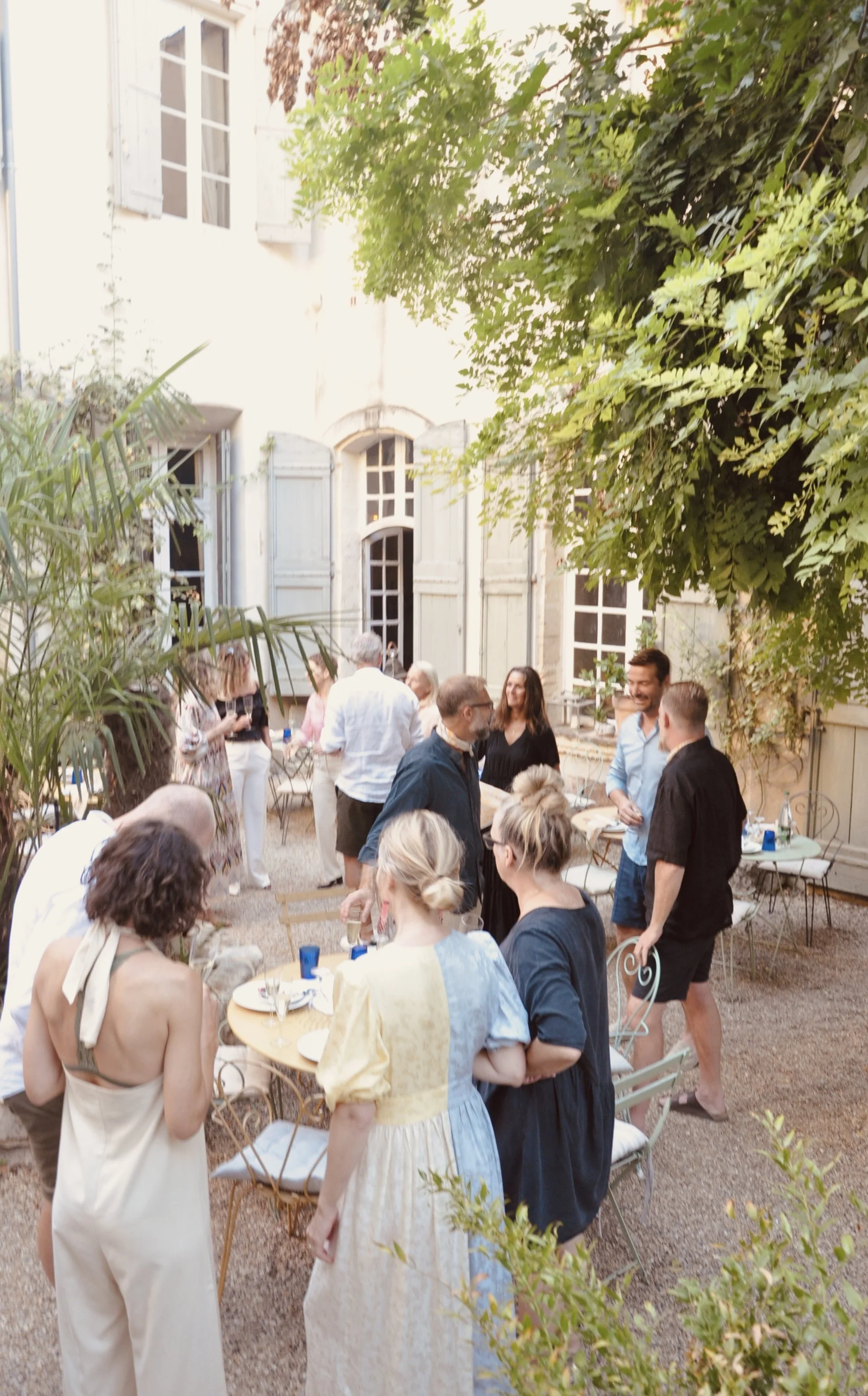 People gathered outdoors in a garden courtyard, socializing and dining during a sunny day. The courtyard is surrounded by a white building with large windows and light blue shutters, with greenery and trees providing shade.
