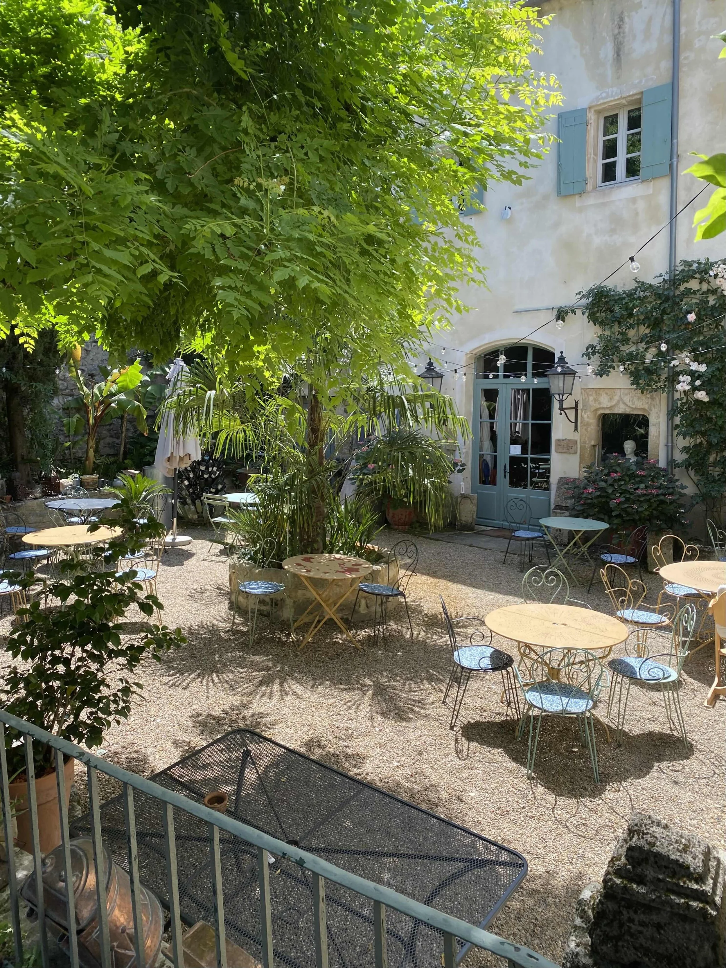Outdoor patio with round tables, metal chairs, lush greenery, a large tree, and a stone building with blue shutters.