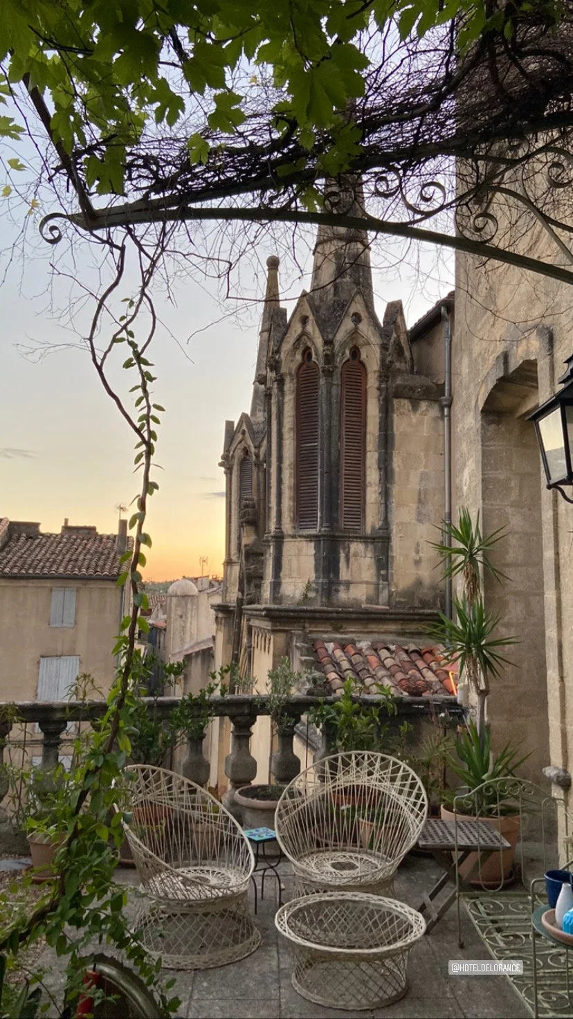 A cozy rooftop terrace with outdoor wicker chairs, potted plants, and a view of a historic stone church with arched windows at sunset.