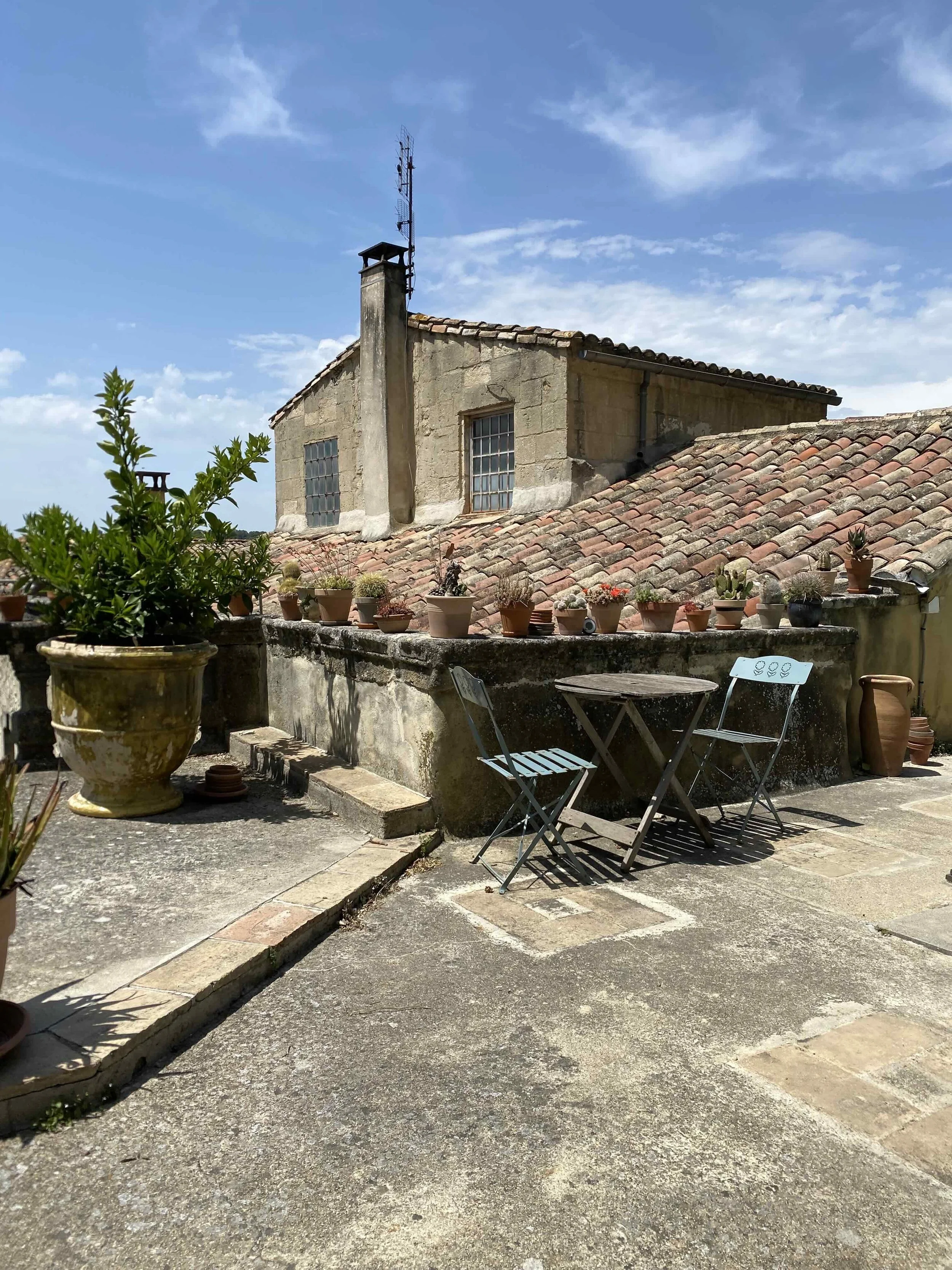 Rooftop terrace with potted plants, a small table, and two chairs, overlooking an older stone building with a tiled roof and a blue sky.