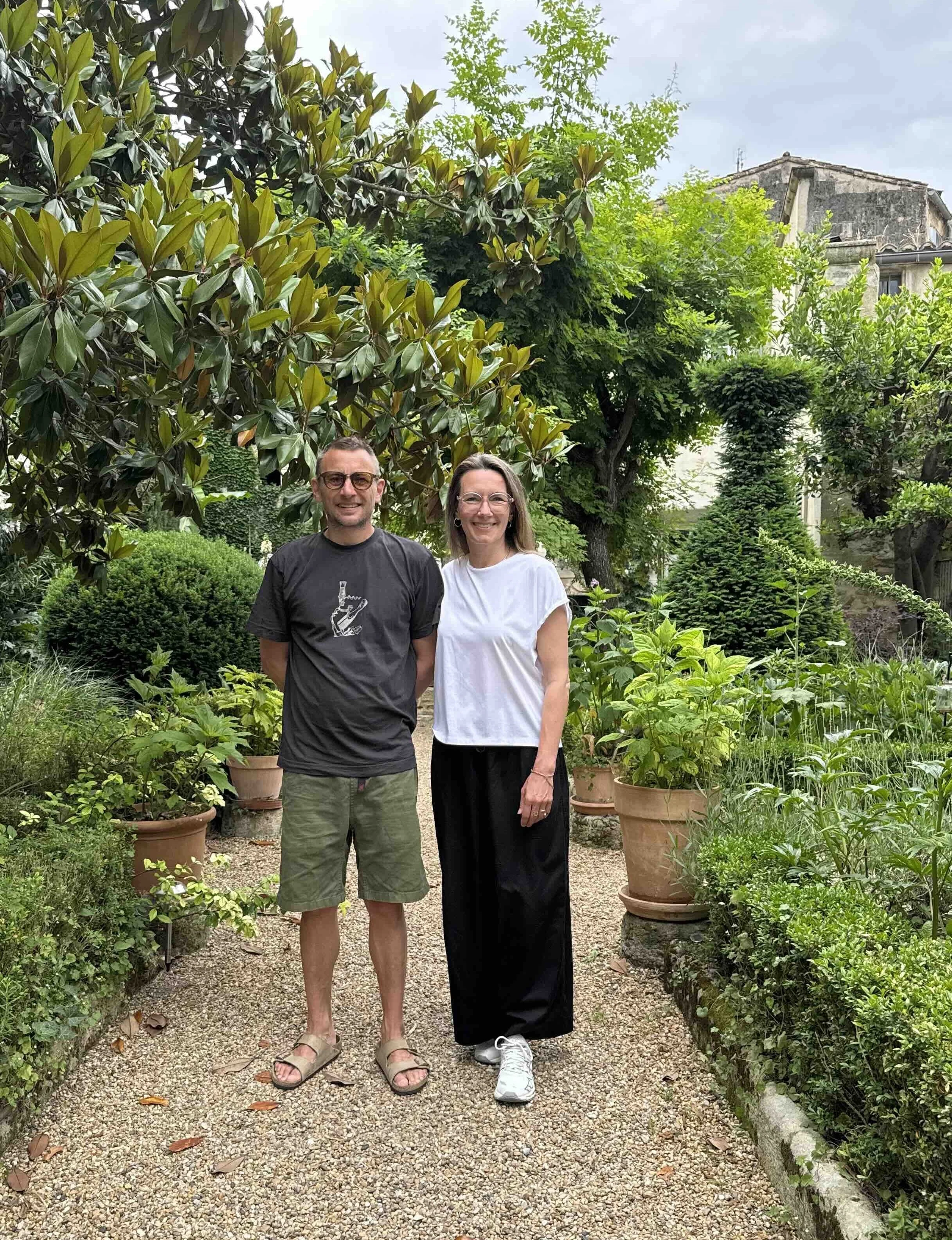Man and woman standing in a lush garden with various green plants and trees, smiling at the camera.