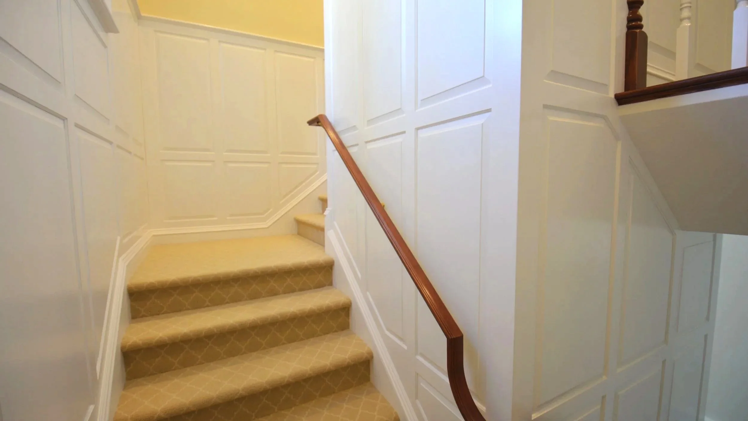 Interior staircase with beige patterned carpet, cream-colored wainscoting walls, and wooden handrails.