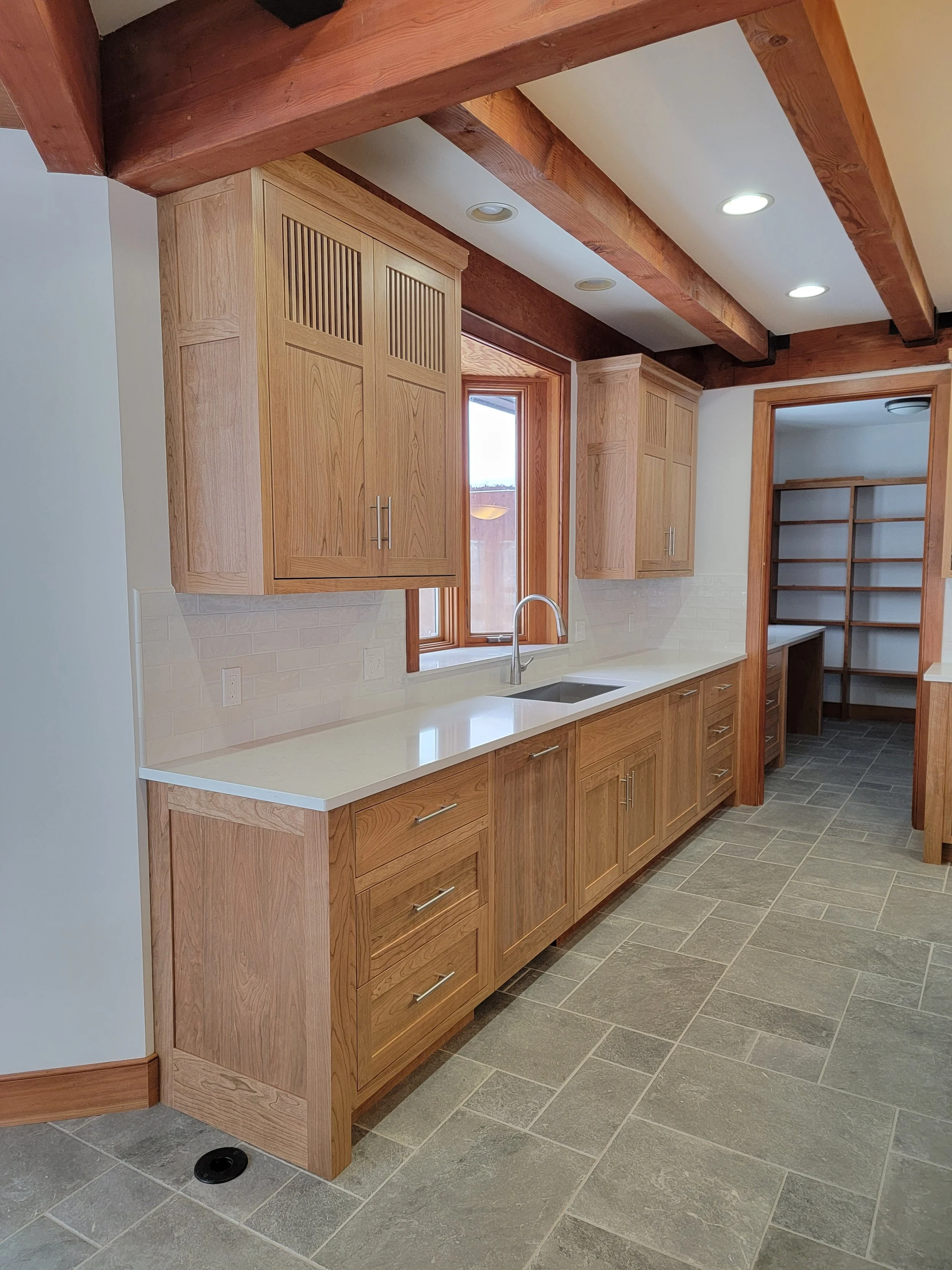 Kitchen with wooden cabinets, white countertop, stainless steel sink, and window above the sink. Wooden beams on ceiling, tiled floor, and open pantry with shelves in the background.