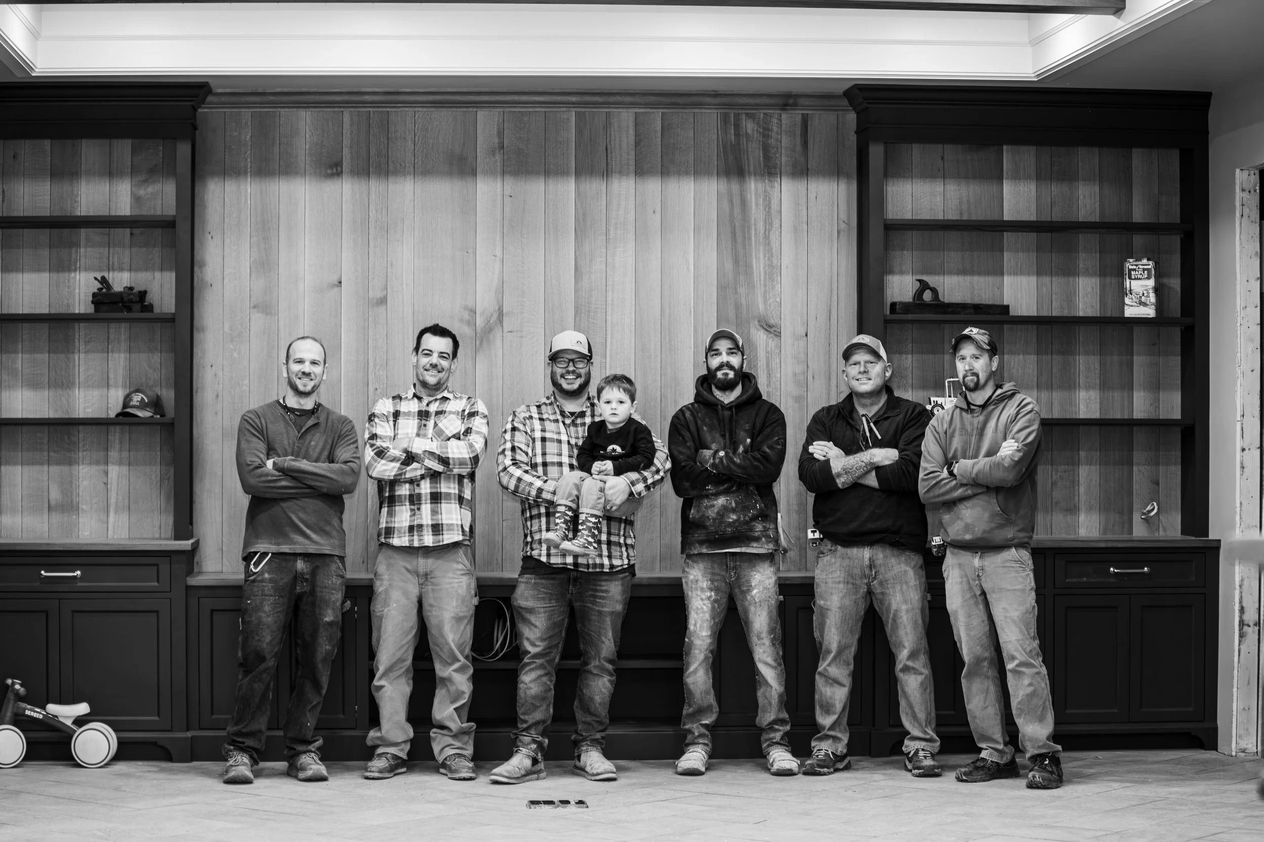 Group of seven men and one child standing indoors in front of a wooden wall with shelves on both sides, smiling with arms crossed, black and white photo.