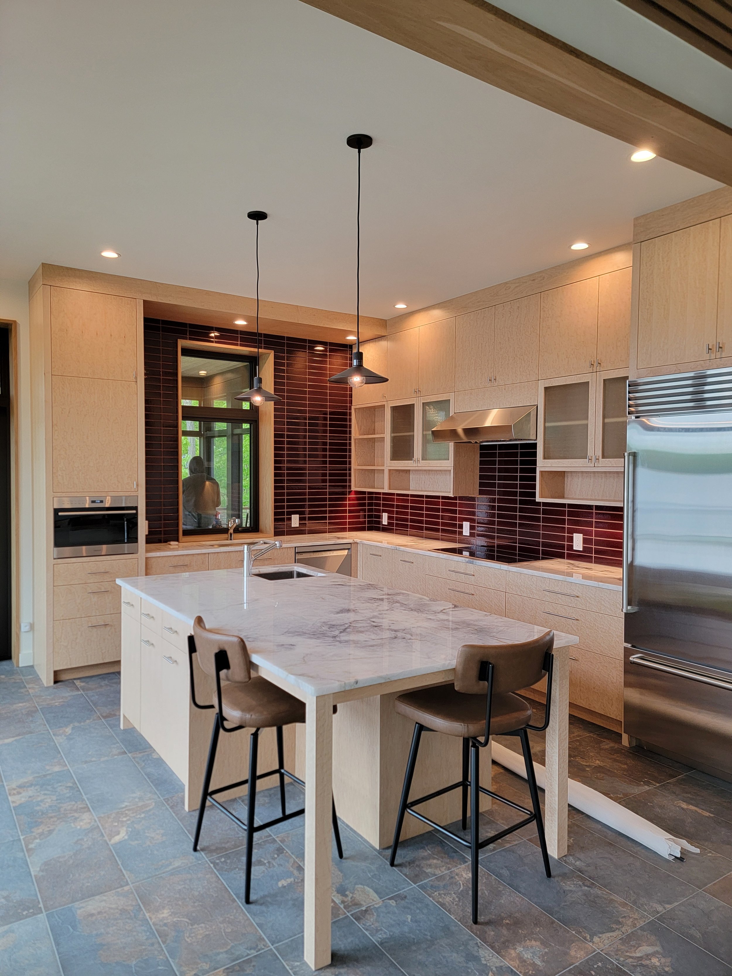 Modern kitchen featuring custom birdseye maple cabinetry with clean lines and minimalist detailing. The natural figuring of the maple highlights the sleek contemporary design while large windows bring natural light and lake views into the space.