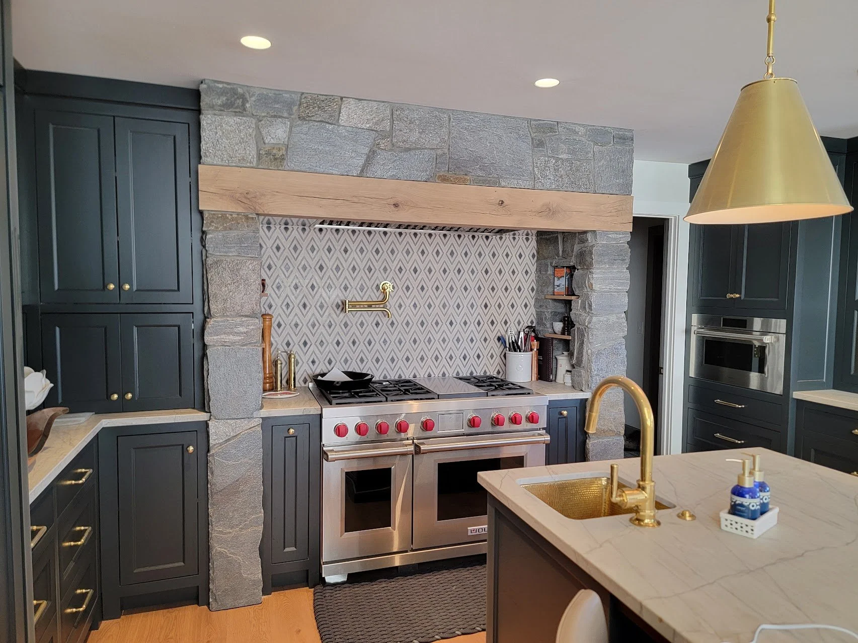 Modern kitchen with dark blue cabinets, stainless steel oven with red knobs, a marble countertop island with gold faucet, and a stone and wood fireplace with patterned tile behind the stove.