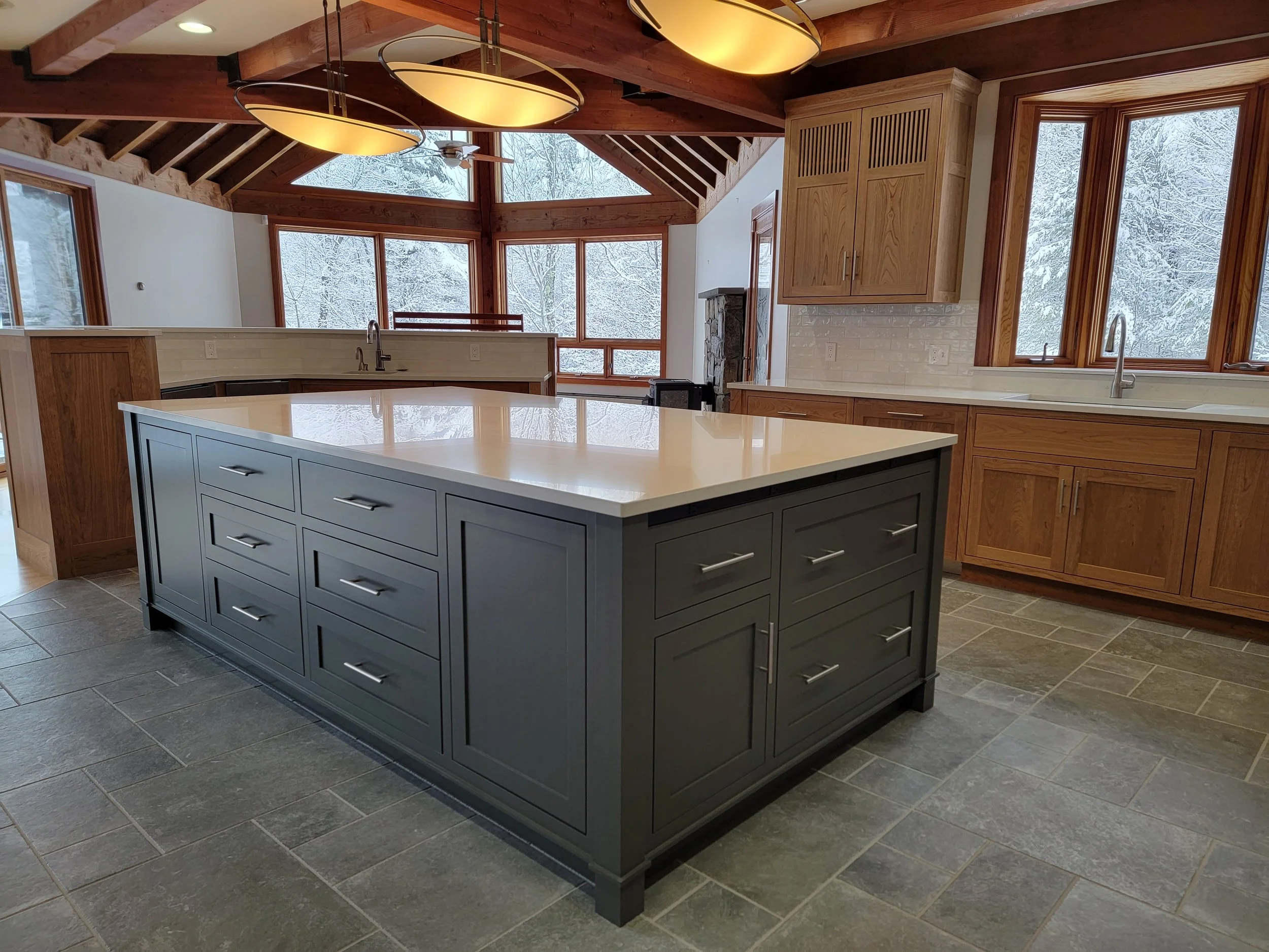 Kitchen with a large navy blue island with a white countertop, wooden cabinets, and large windows showing a snowy landscape outside.