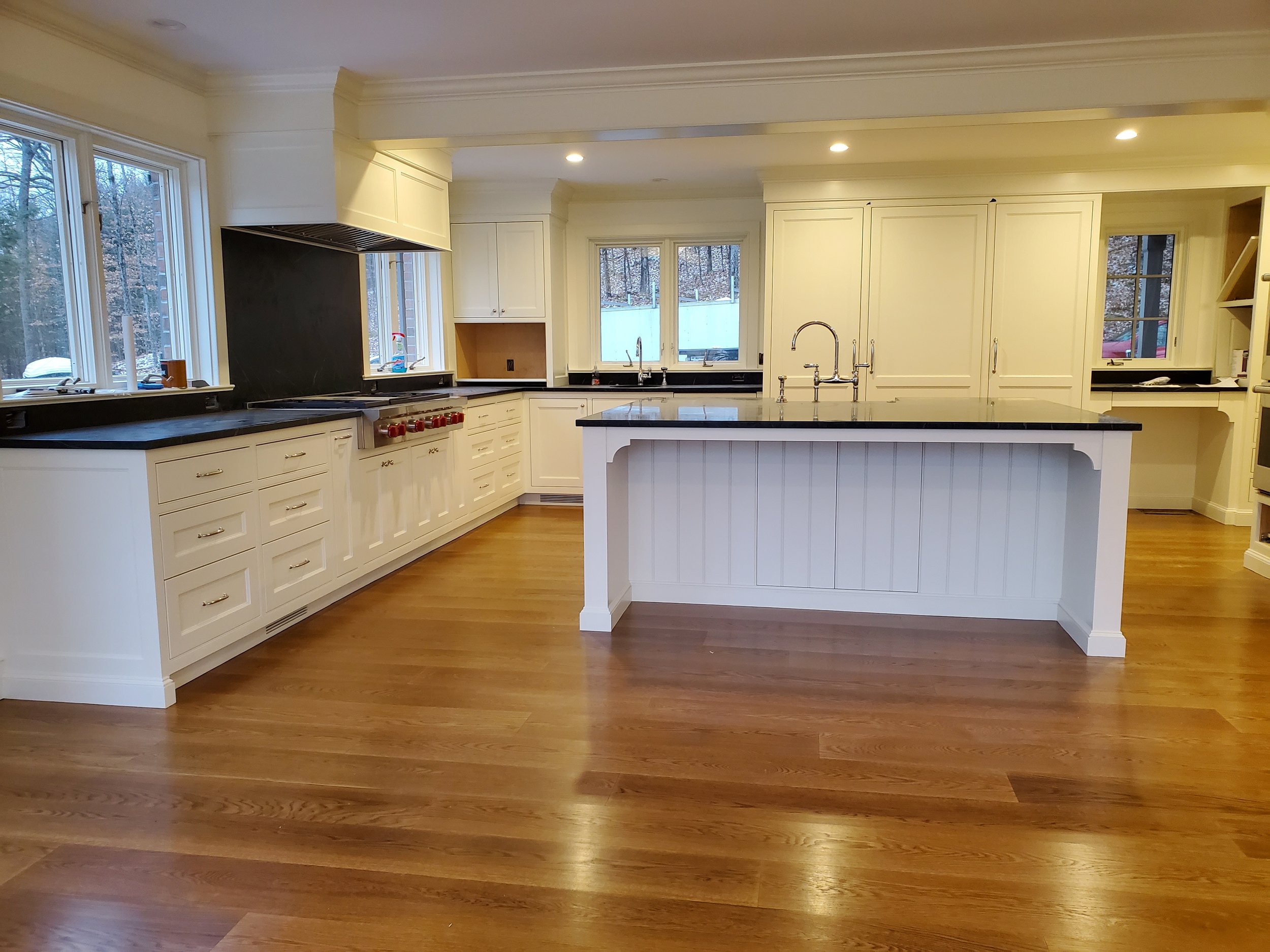 Modern kitchen with white cabinetry, black countertops, central island with a double sink, hardwood floors, and multiple windows overlooking a wooded yard.