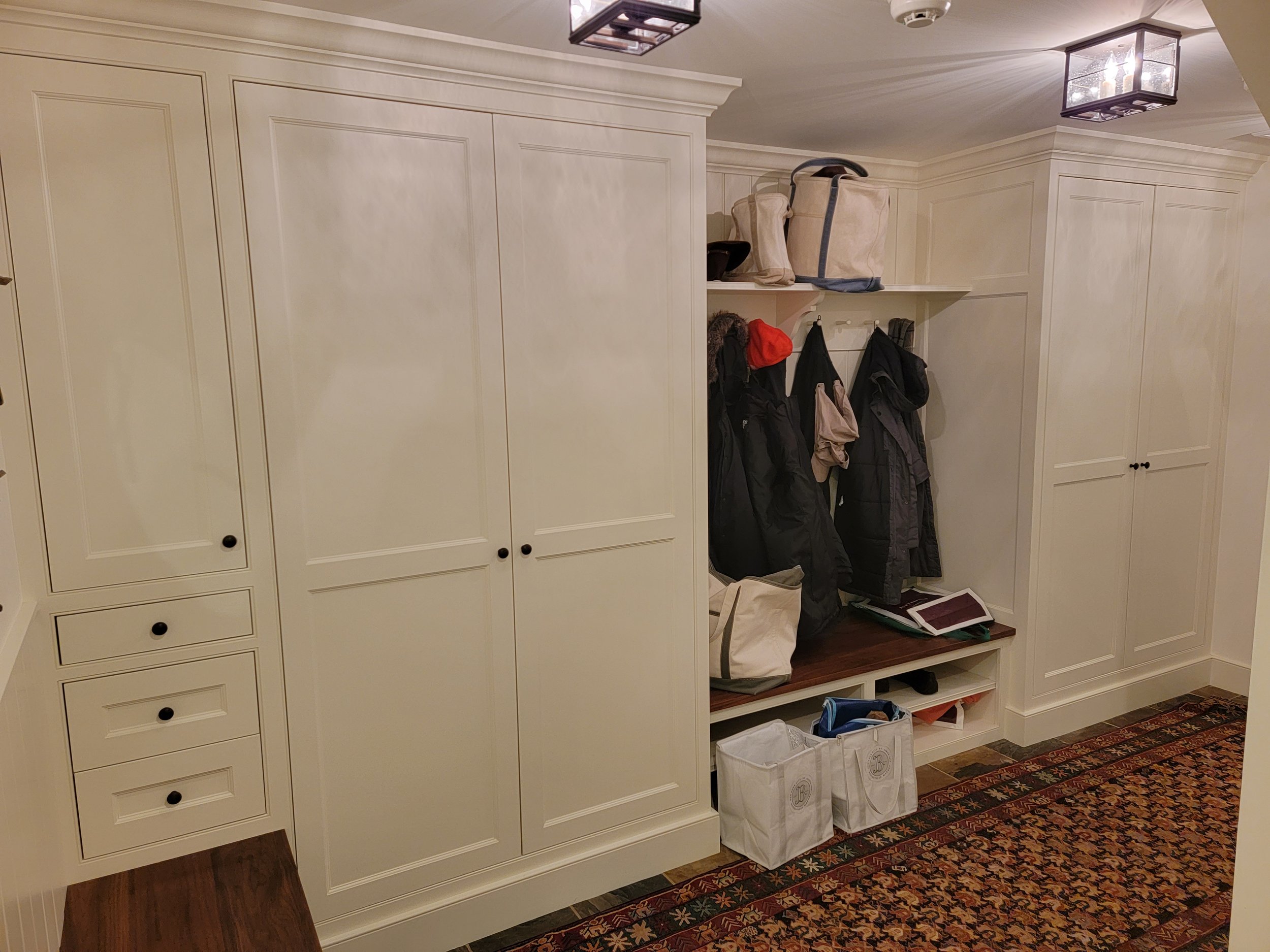 Entryway with white built-in cabinets and coat hooks, containing bags, jackets, and hats, with a multicolored rug on the floor and ceiling lights.