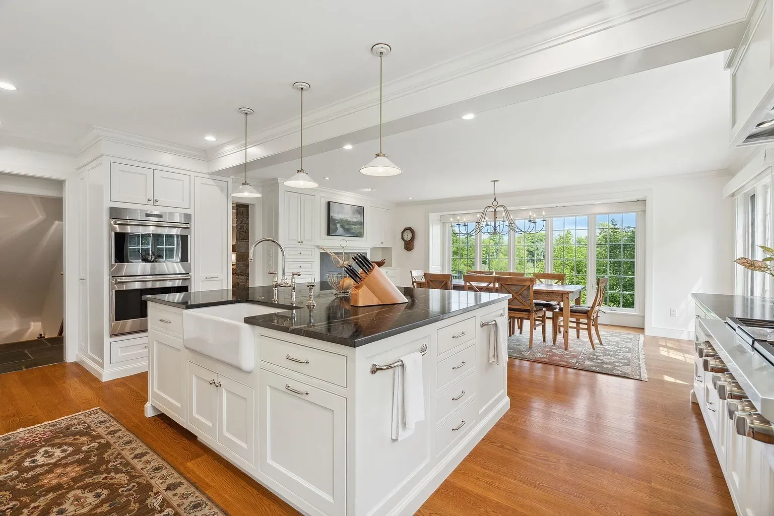 Bright, spacious kitchen with white cabinets, a large island with a dark countertop, stainless steel appliances, and a dining area with large windows showing green trees outside.