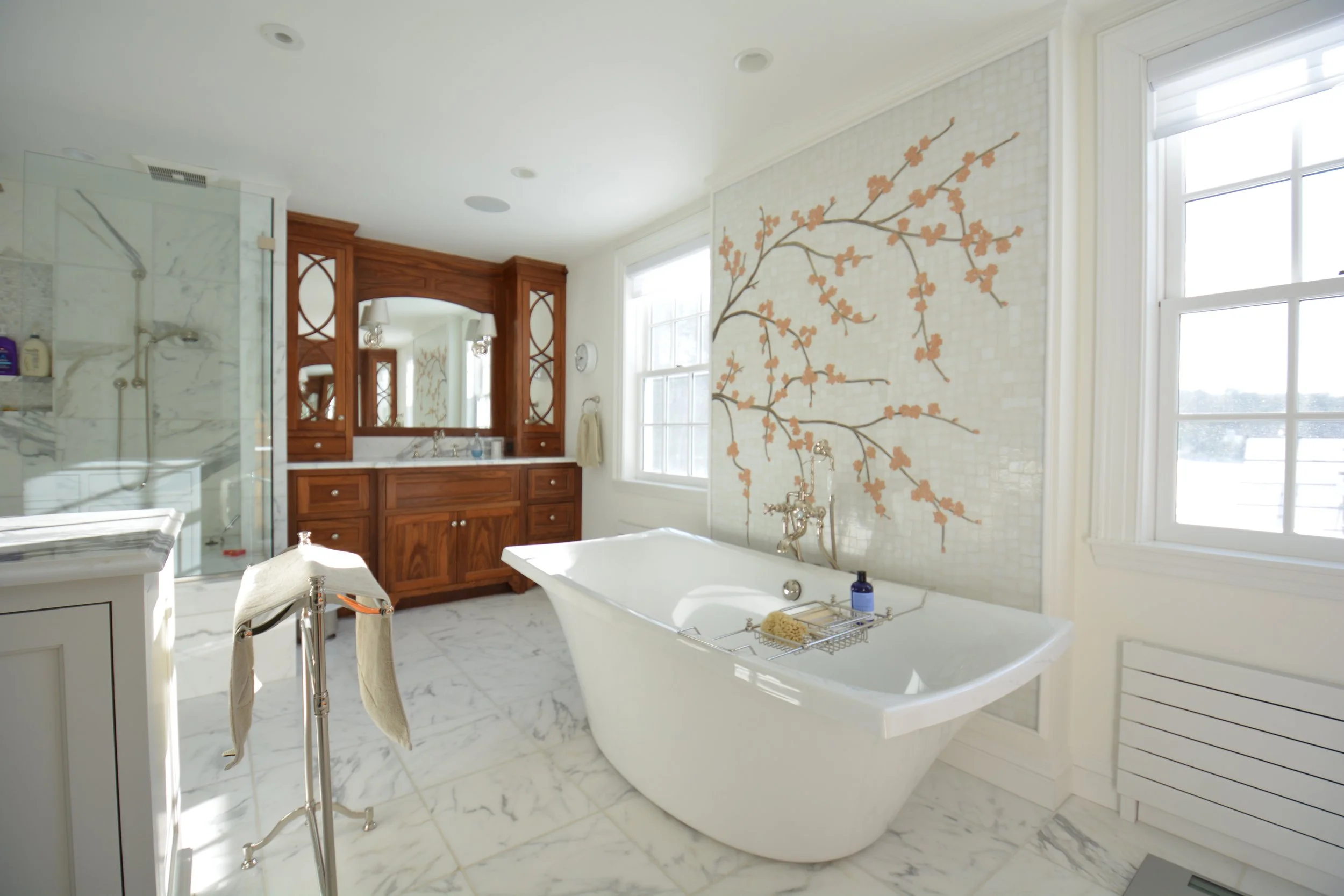 Bright bathroom with a freestanding bathtub, wooden vanity with mirror, marble floor and wall, floral wall art, large windows, and glass-enclosed shower.