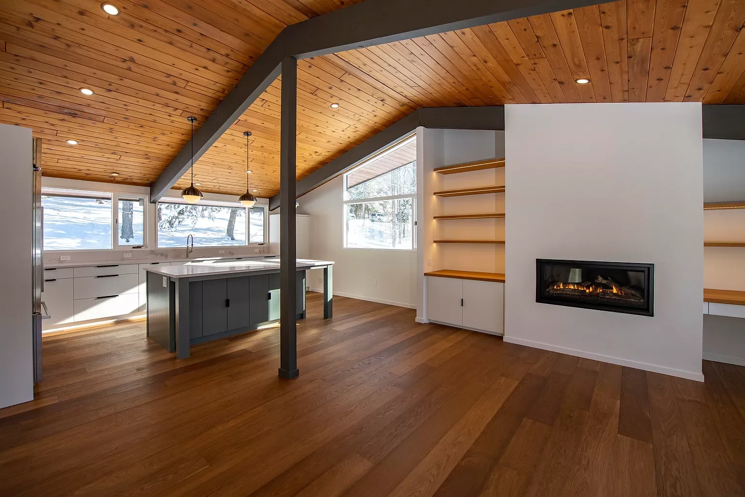 Modern kitchen and living area with wooden ceiling, hardwood floors, built-in shelves, a gas fireplace, and large windows with a snowy outdoor view.