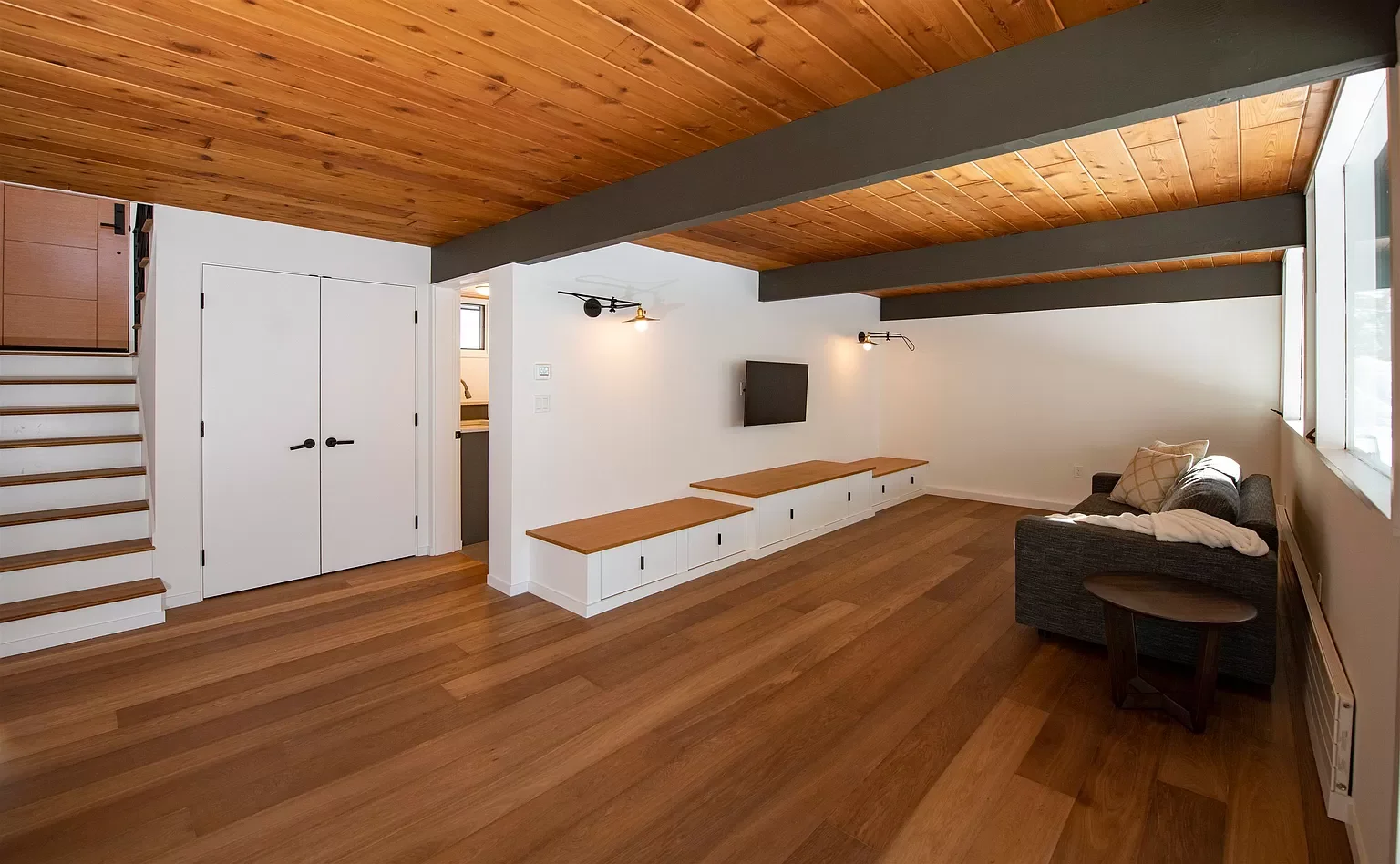 Living room with wooden floors and ceiling, white walls, a gray couch with pillows, a wall-mounted TV, small wooden side table, and built-in white storage along one wall. Large window letting in natural light.