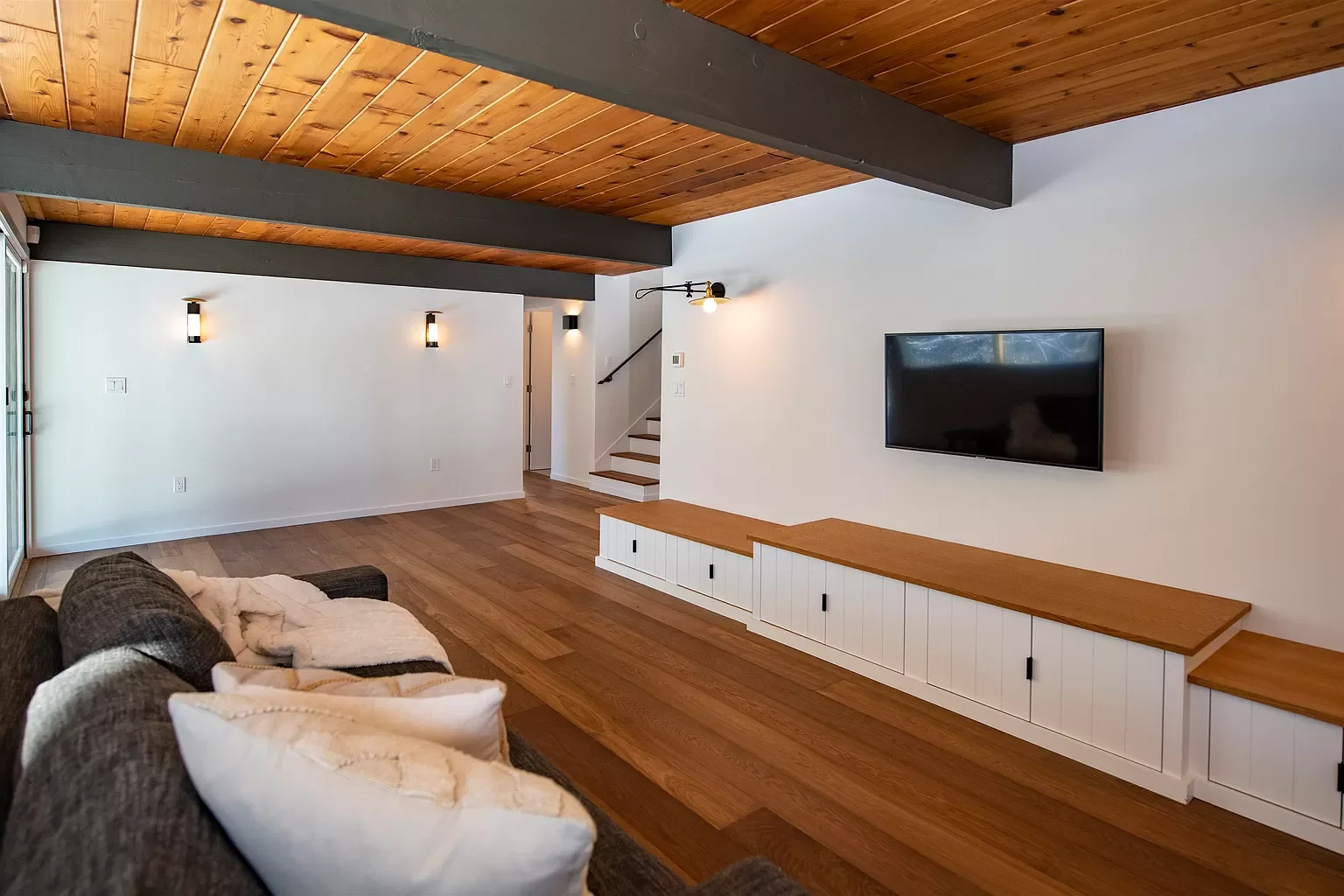 Modern living room with a wooden ceiling, wall-mounted TV, built-in storage, a leather sofa with cushions, and wall-mounted lights.