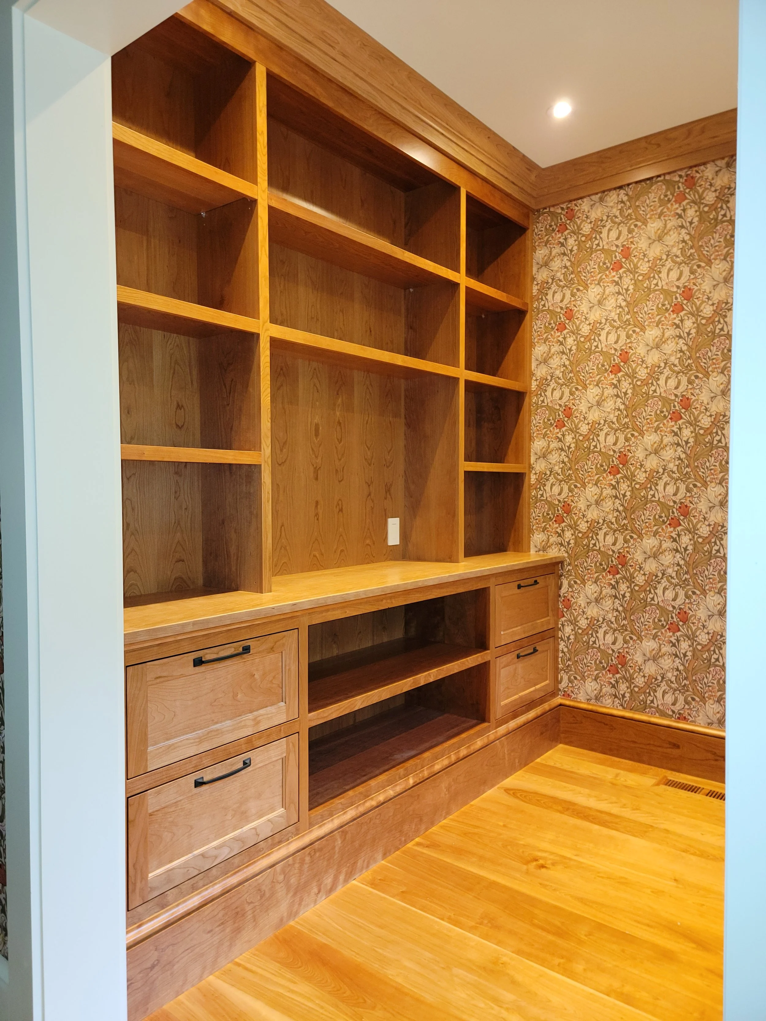 Empty built-in wooden bookshelf with multiple open shelves and drawers in a room with wood flooring and floral wallpaper.