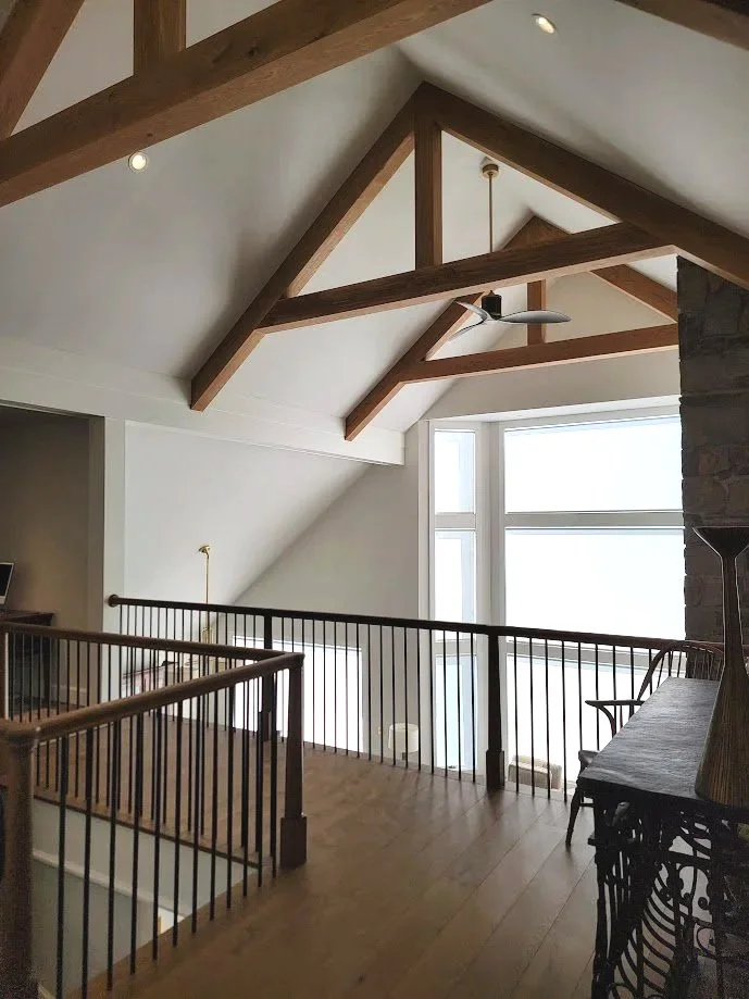 Interior view of a room with large windows, exposed wooden beams on the ceiling, and a wooden floor. There is a black metal railing along the staircase opening and a wooden bench or furniture near the window.