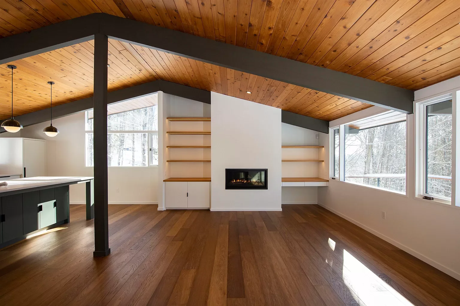 Empty modern living room with large windows, wooden ceiling, hardwood floors, a fireplace built into a white wall, and built-in shelves with natural wood trim.