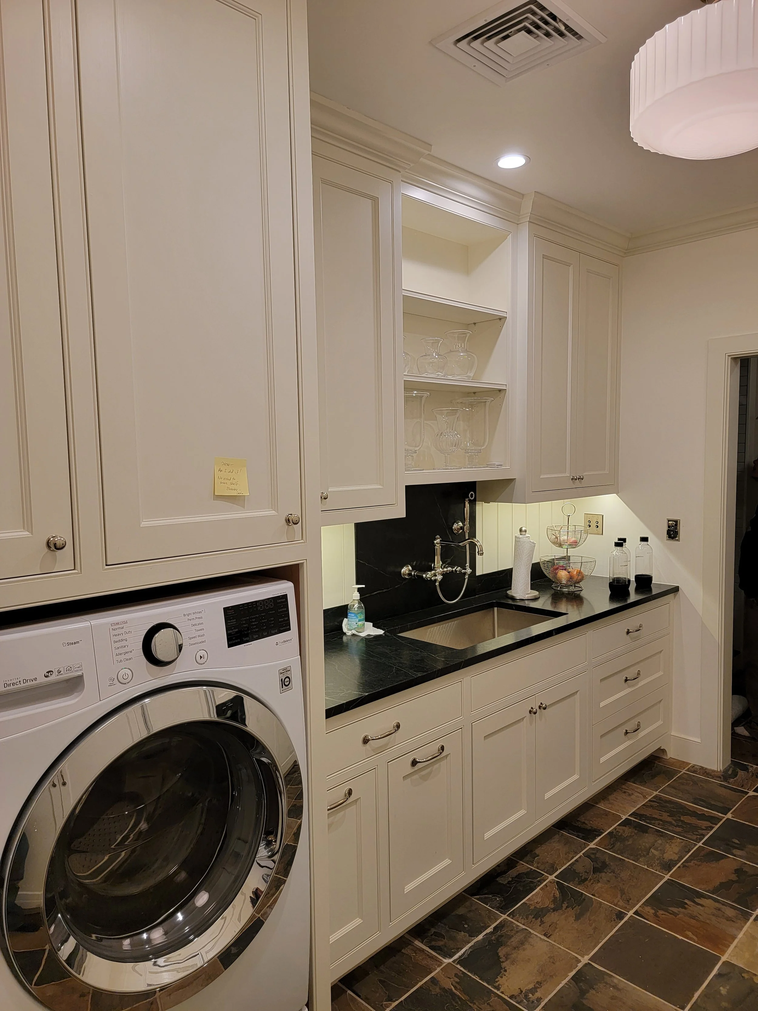A laundry room with white cabinetry, black countertop, and a front-loading washing machine. There are open shelves with glassware, a small sink, and various containers and bottles on the countertop.