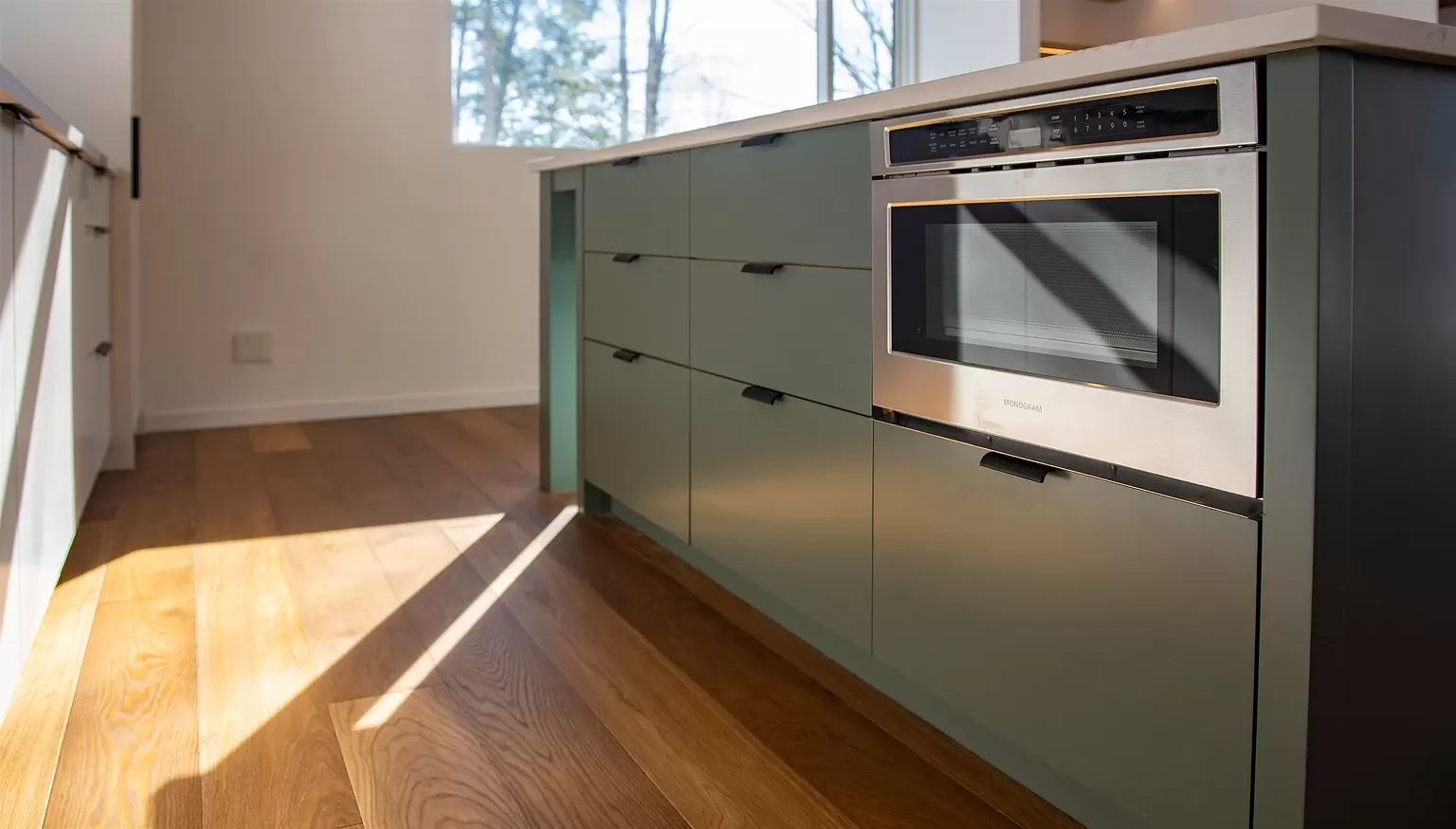 Modern kitchen with green cabinetry, stainless steel oven, and hardwood floor, with sunlight streaming through a window.