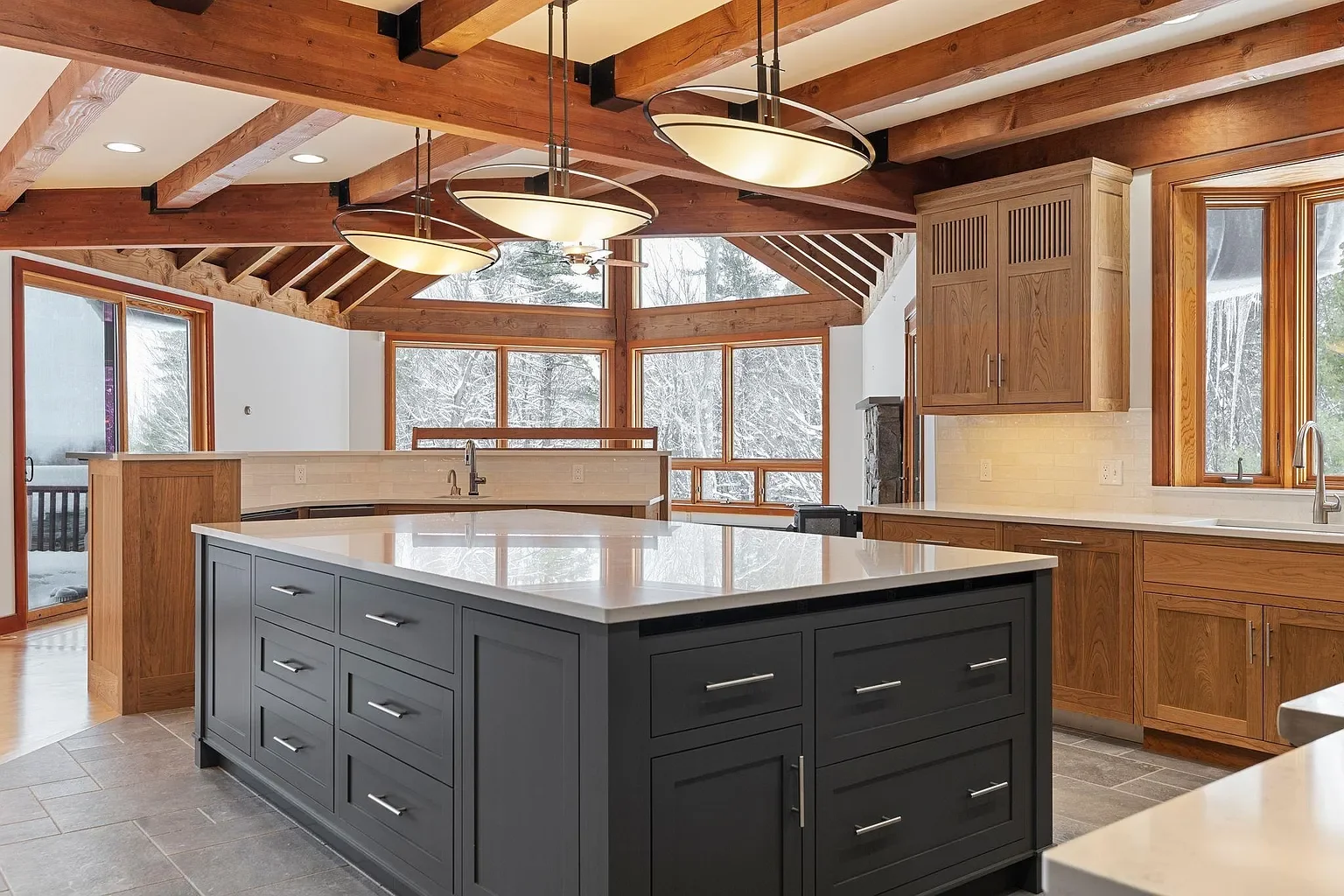 Modern kitchen with wooden beams on the ceiling, a large gray island with drawers, wooden cabinets, and large windows showing a snowy outdoor scene.