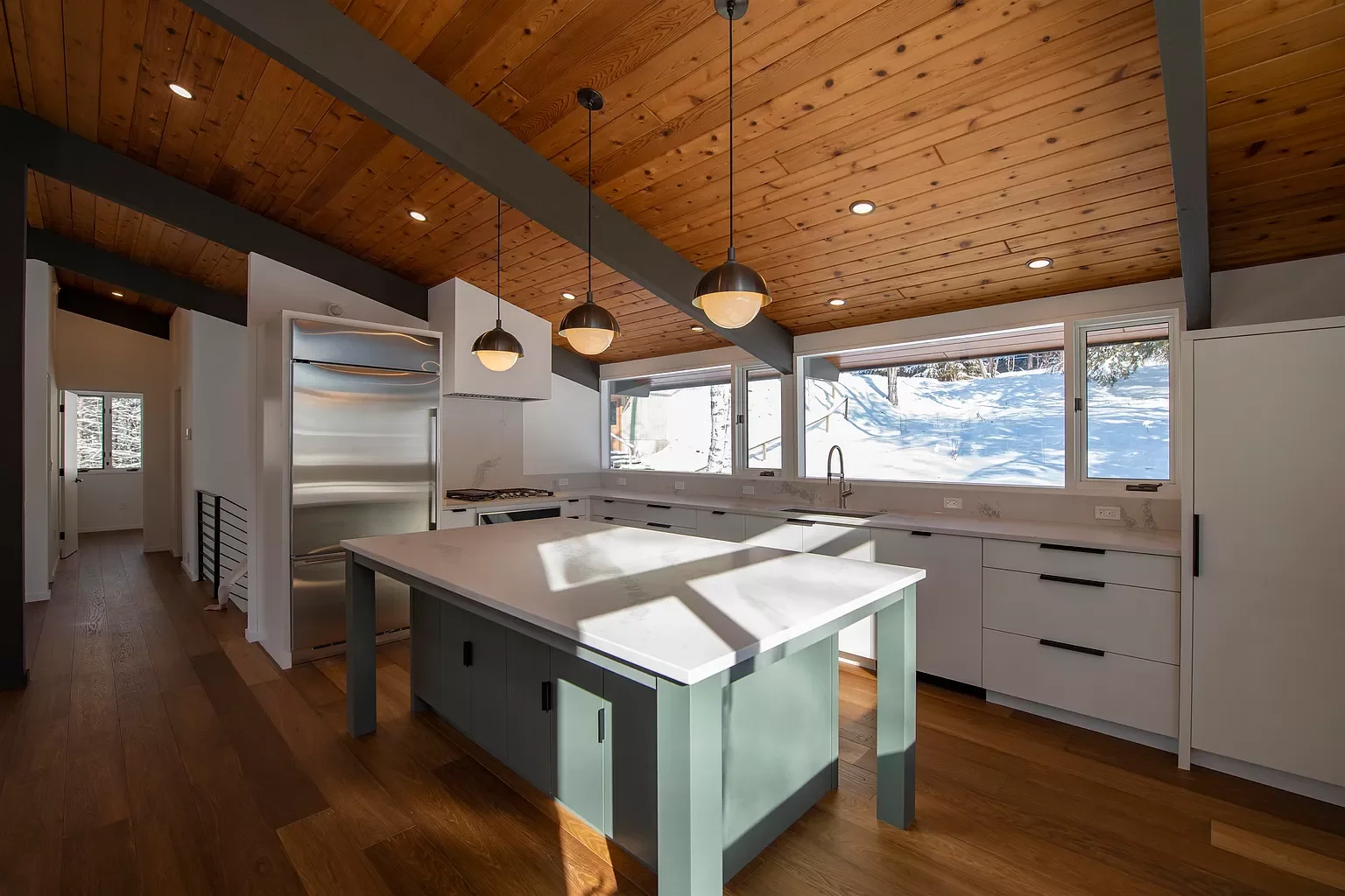 Modern kitchen with white cabinets, a kitchen island with a pale blue base, a wooden ceiling, and large windows showing a snowy landscape outside.