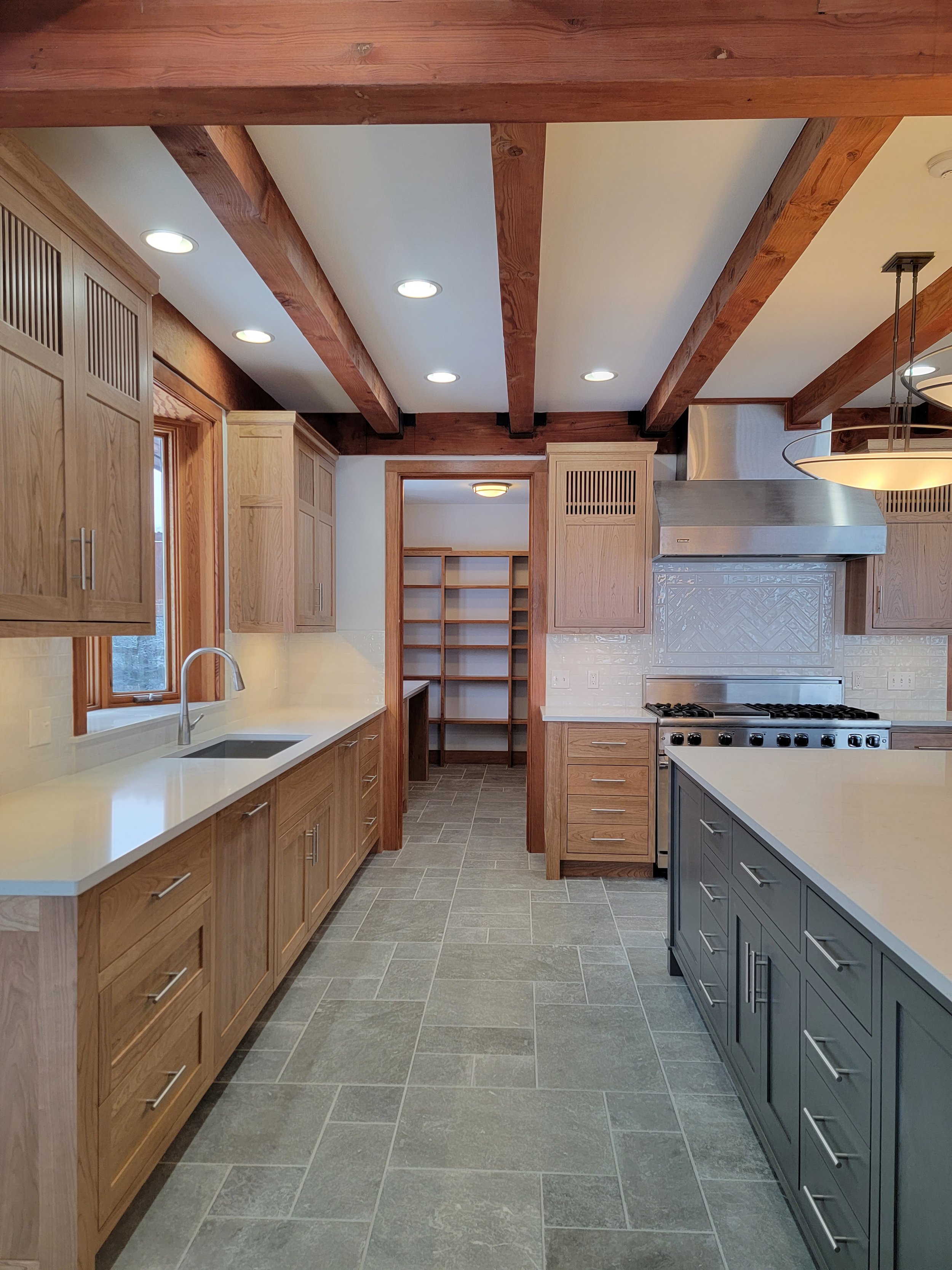 Kitchen with wooden cabinets, a window above the sink, gray tiled floor, and a large stove with a metal hood. A pantry with open shelves is visible in the background.