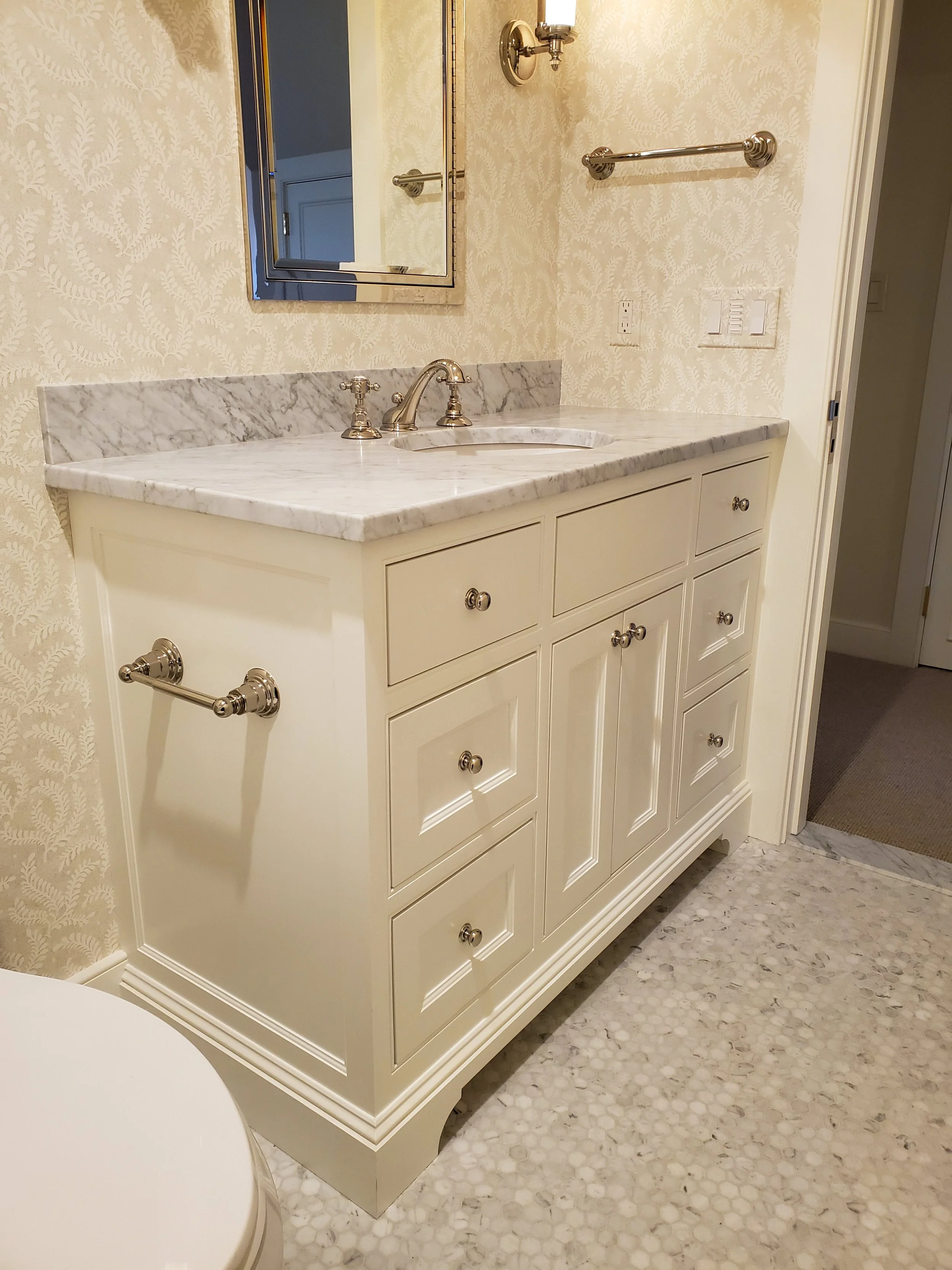 A cream-colored bathroom vanity with a marble countertop, silver knobs, and a central faucet. There is a mirror above the sink, a wall-mounted light fixture, and a towel bar. The wallpaper has a light cream and beige leafy pattern, and the floor is c