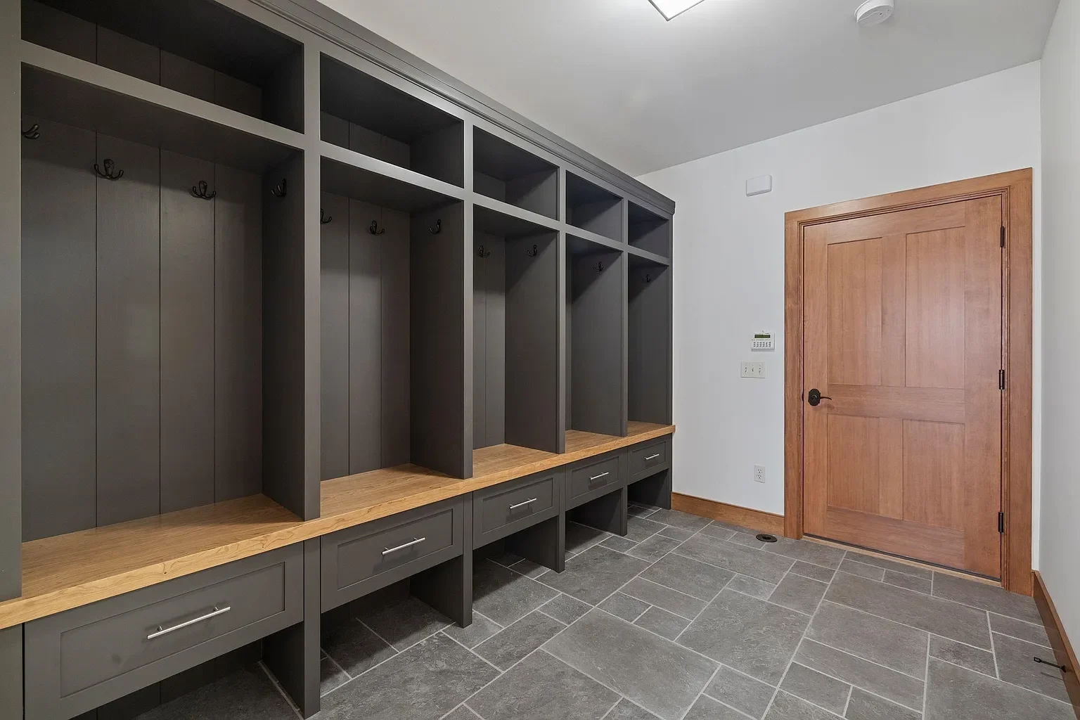 A modern mudroom with gray storage cubbies and hooks, a wooden countertop, and a wooden door. The room has gray tiled flooring and white walls.