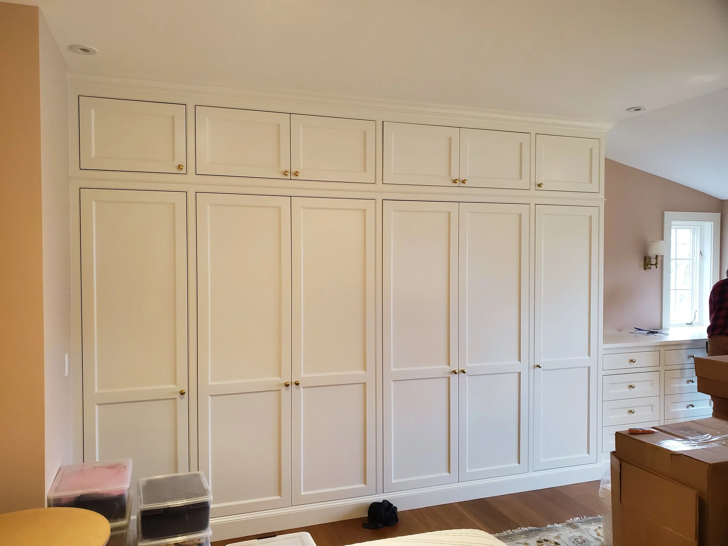 A large white built-in closet with multiple doors and small gold knobs in a room with wooden floors and a beige wall, featuring a small window and a person with red plaid shirt partially visible.