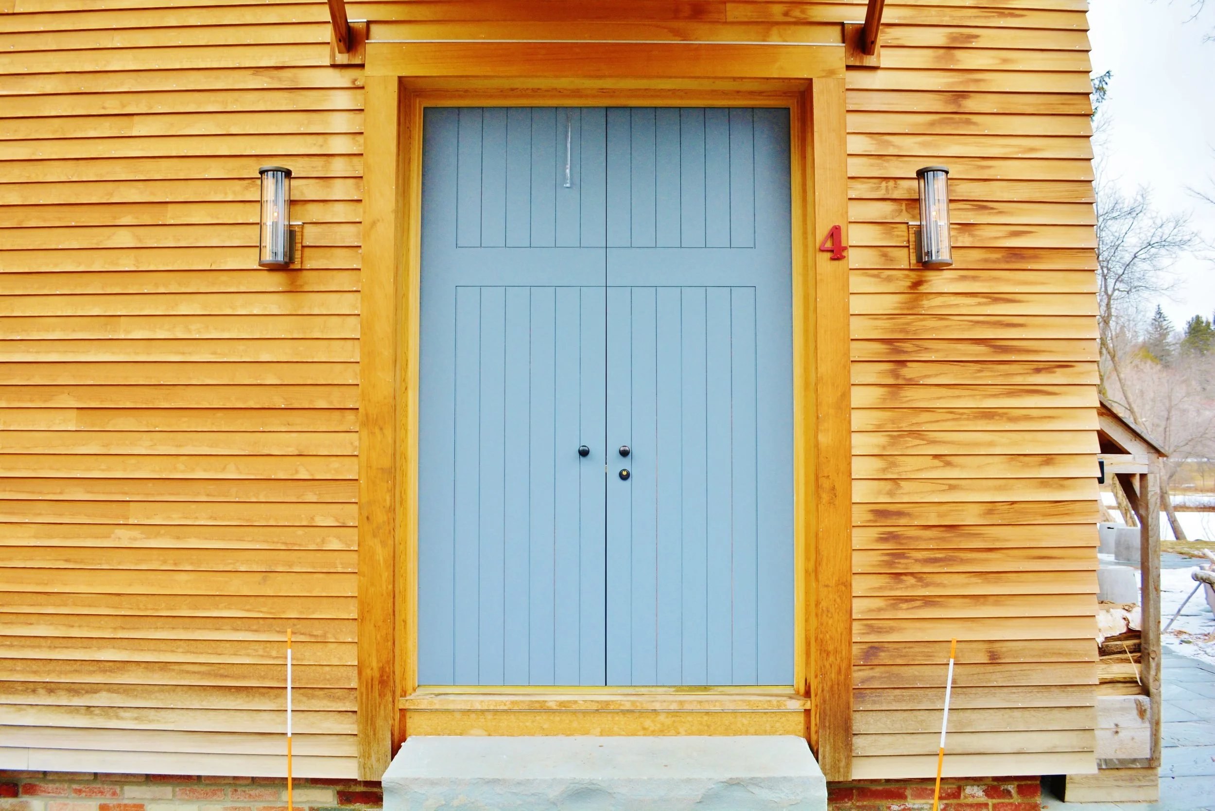 Front view of a wooden house with a blue door, two wall-mounted lights on each side, a small concrete step in front, and the number 4 in red on the right side of the door.