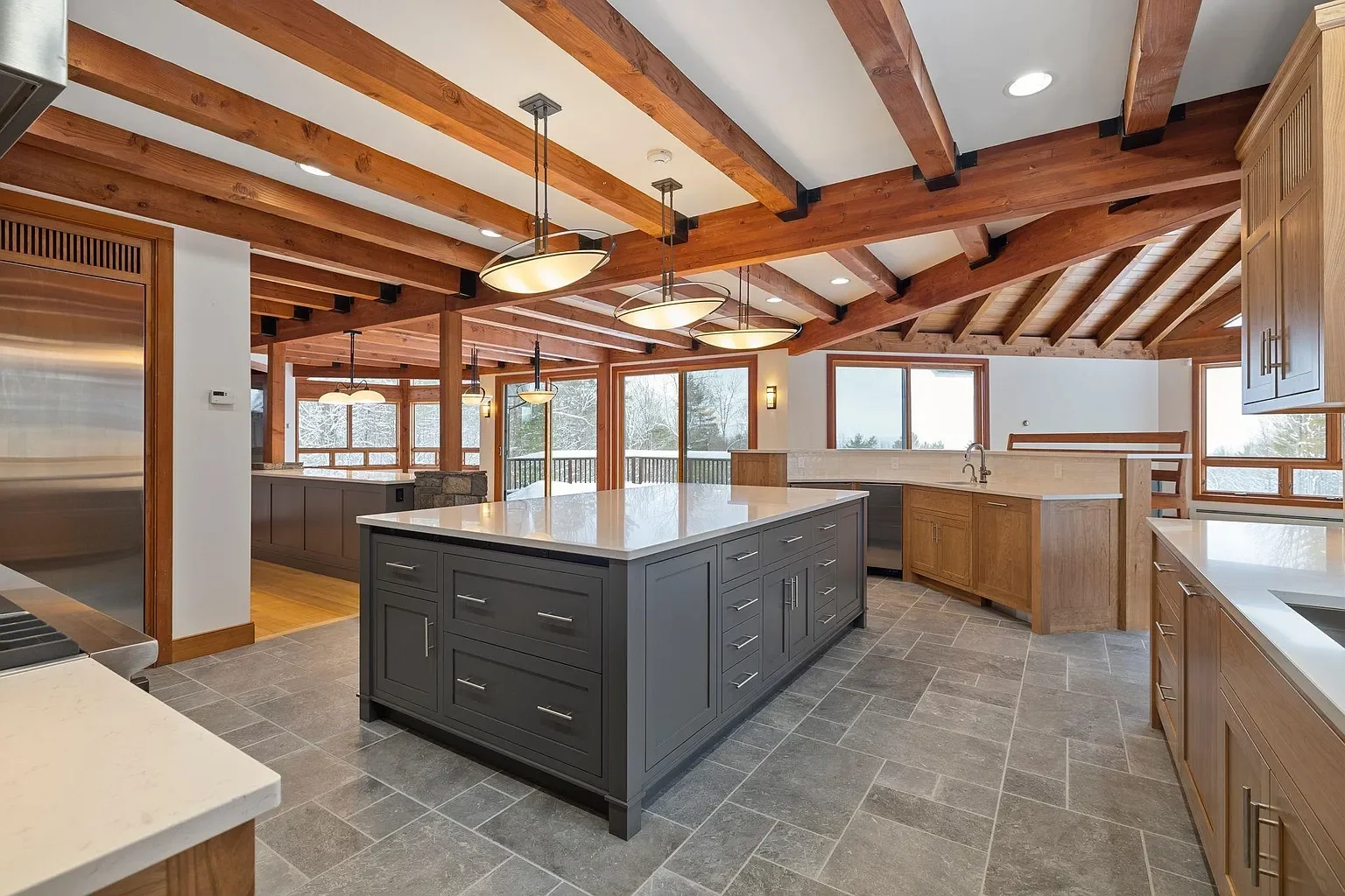 Modern kitchen with wooden beams, large island with gray cabinets, and ample windows showing a snowy outdoor landscape.