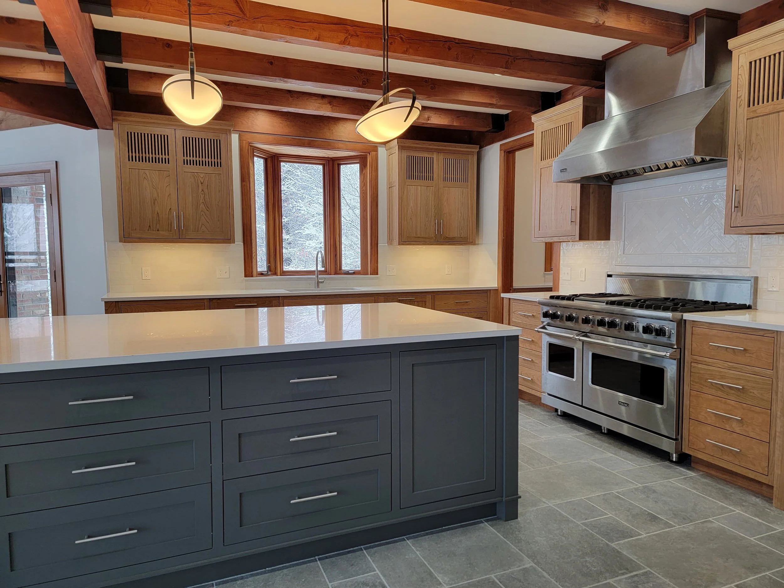Modern kitchen with wooden cabinets, a gray island with drawers, stainless steel oven and range, range hood, and a window with snow outside.