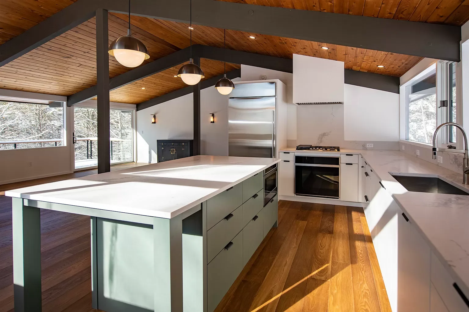 Modern kitchen with a white marble countertop, green island, stainless steel refrigerator, built-in oven, gas stove, and large windows showing snow-covered trees outside.