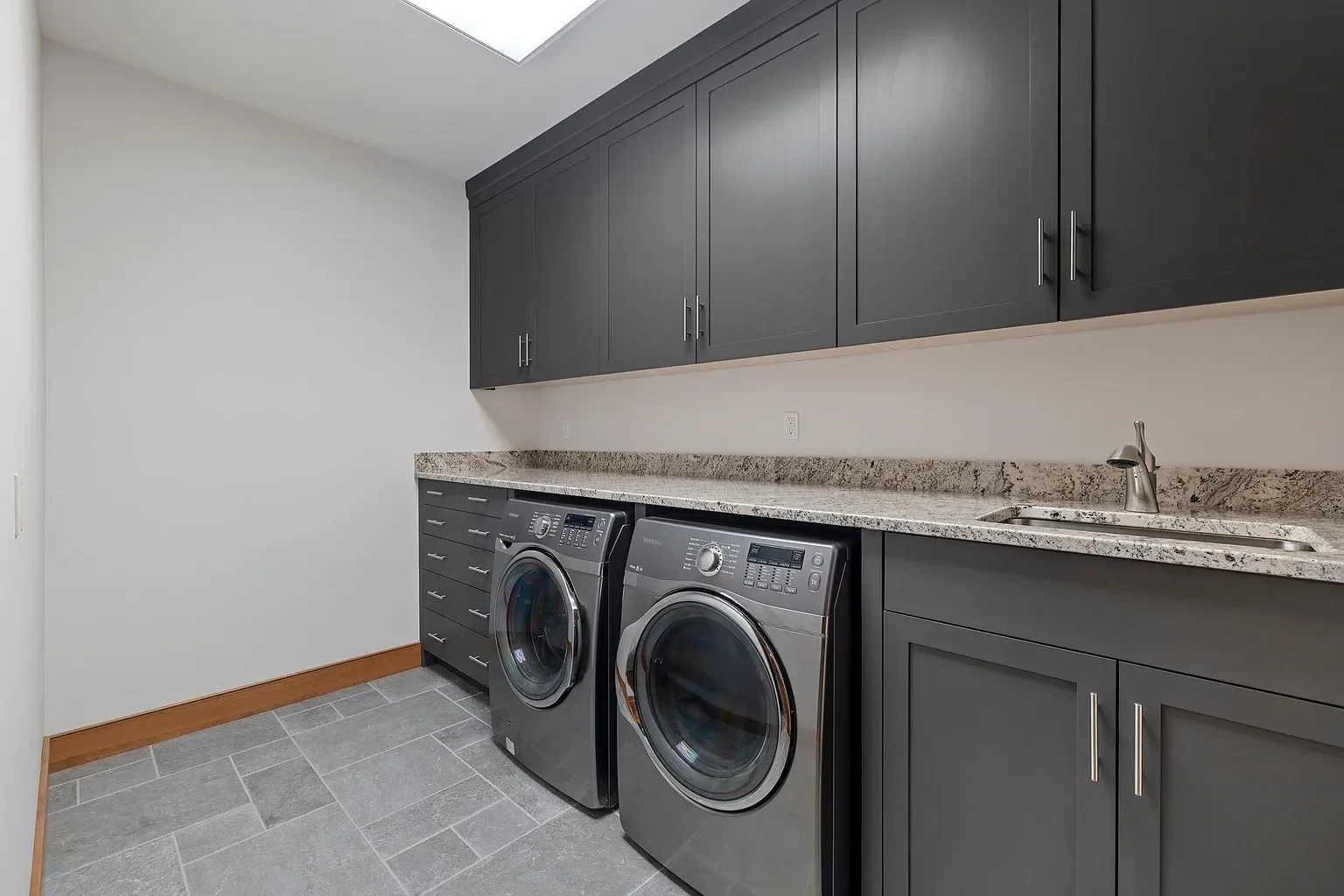 A laundry room with grey cabinets, a granite countertop, a stainless steel sink, a washing machine, and a dryer. The floor has grey tiles, and there is a white wall with outlets.
