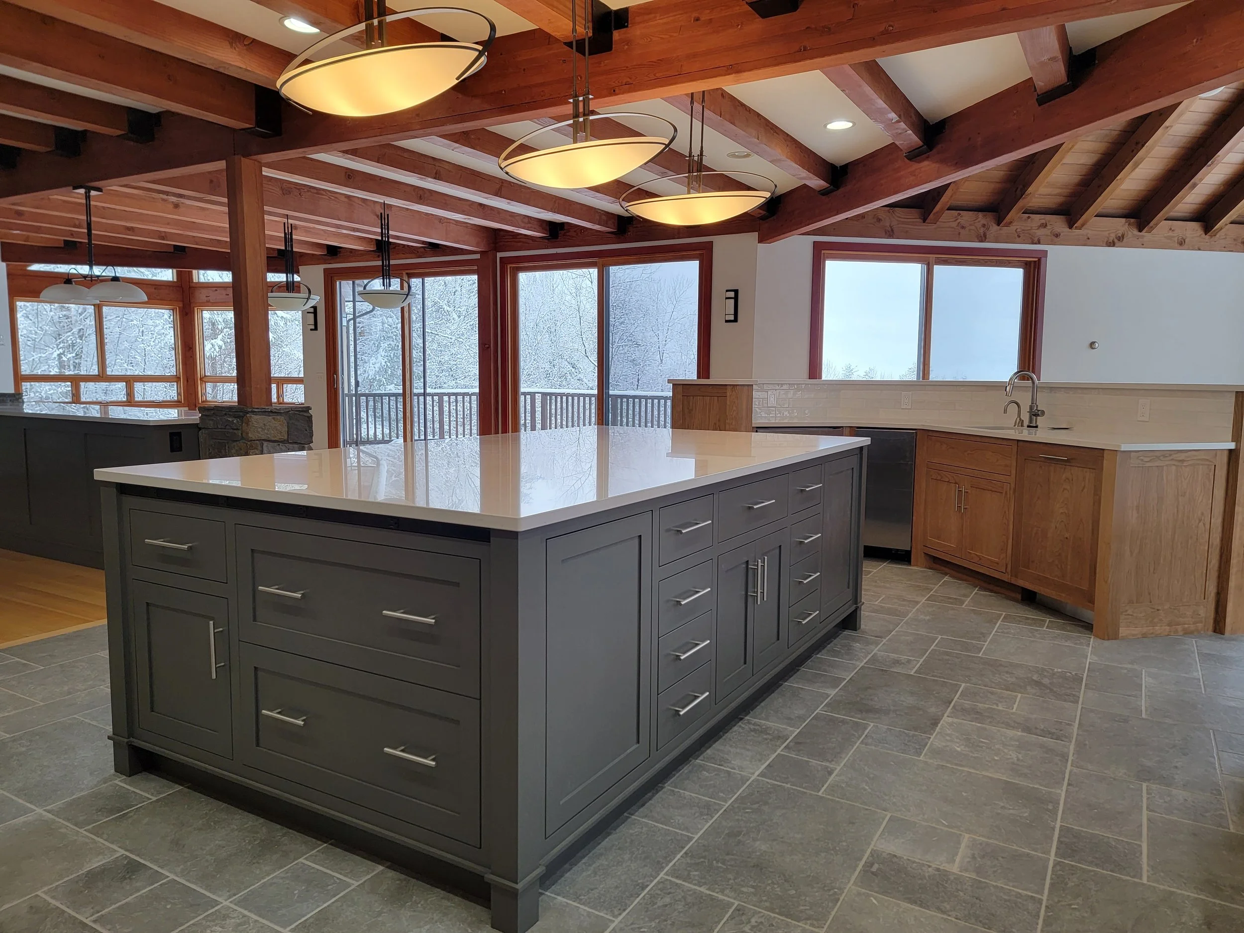 Modern kitchen with gray island, wooden cabinets, large windows overlooking snowy landscape, vaulted wooden ceiling with pendant and chandelier lights, tiled floor.