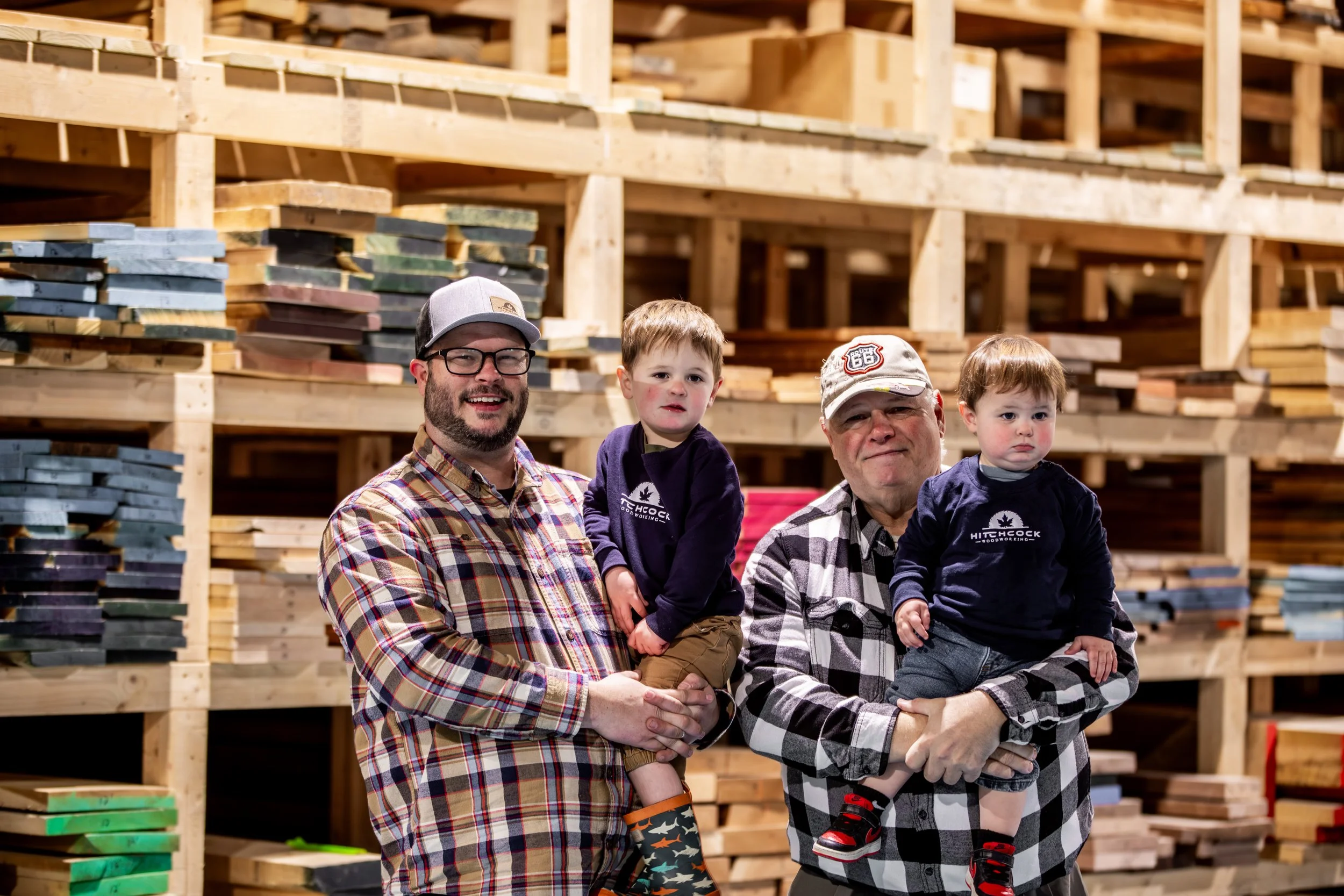 Two men holding young boys inside a lumber warehouse with stacks of wood in the background.