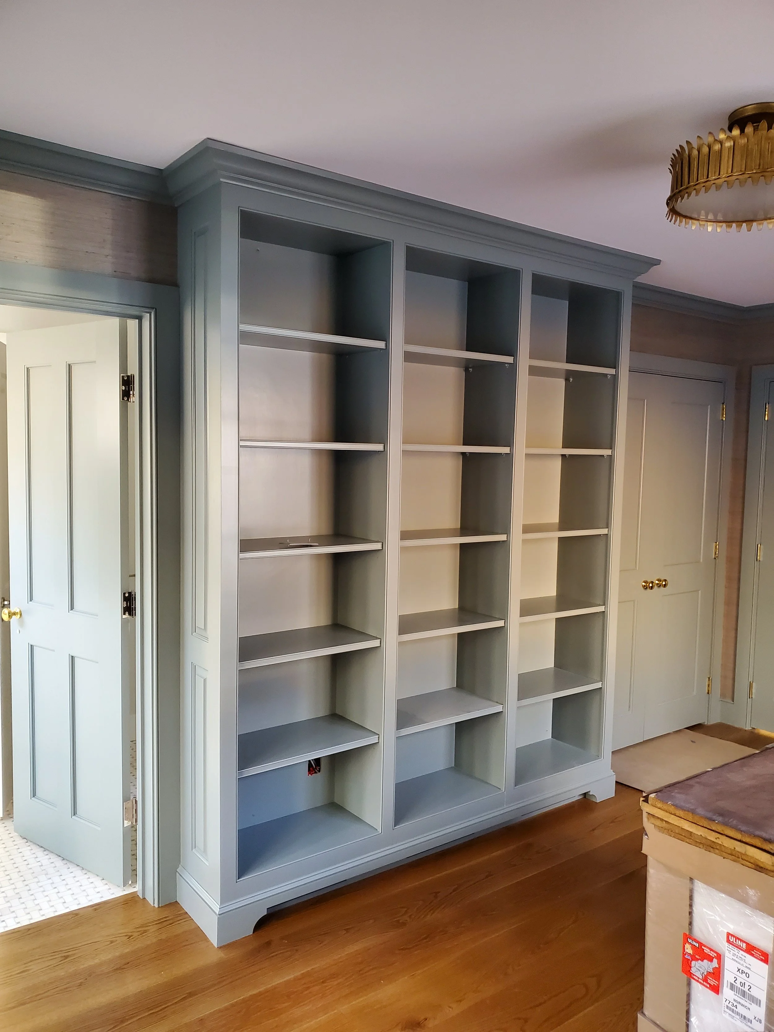 Empty built-in bookshelf with multiple shelves, painted in light gray, located in a room with wooden flooring and light-colored walls.
