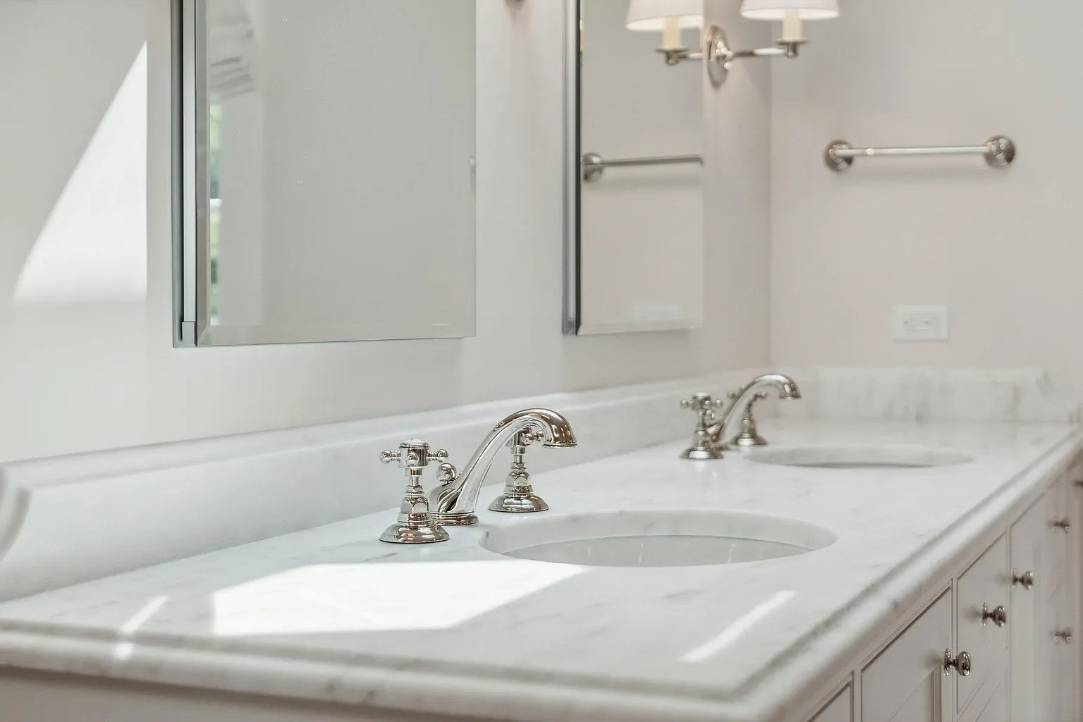 A bathroom double vanity with white marble countertop, two undermount sinks, and chrome faucets. Above the sinks are two mirrors and a wall-mounted cabinet on the left. To the right, there are two towel bars and a wall outlet.