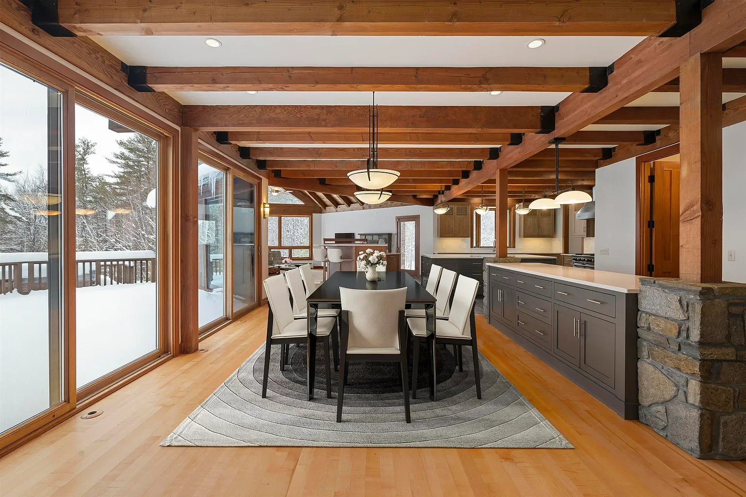 Interior of a dining area and kitchen with large sliding glass doors leading to a snowy outdoor deck, wooden beams on the ceiling, a black dining table with white chairs, and modern pendant lighting.