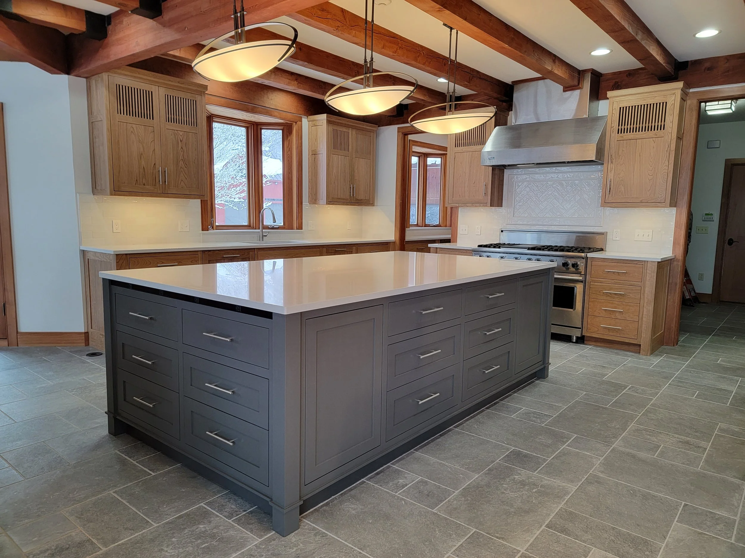 A spacious kitchen with a large center island painted gray with multiple drawers, wooden upper cabinets, and a stainless steel gas stove with a vent hood. The kitchen has wood ceiling beams, pendant and recessed lighting, and a view of snow outside t