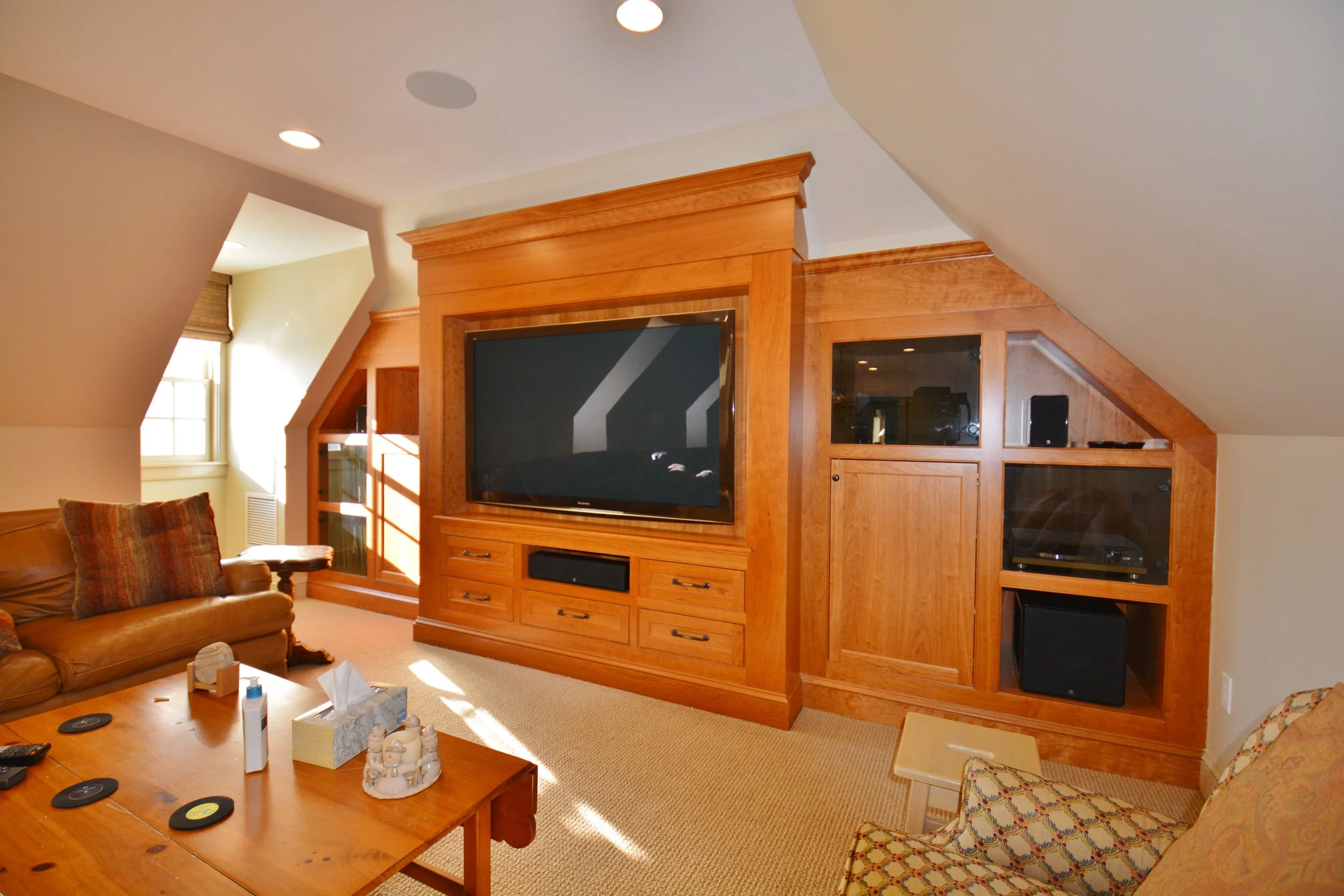 Living room with a large flat-screen TV set in a wooden entertainment center, a leather sofa with pillows, a coffee table, and a patterned armchair. Sunlight from a window illuminates the space.
