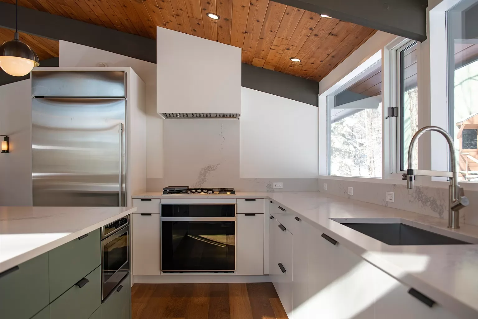 Modern kitchen with white countertops, black handles, stainless steel refrigerator, and large window with a view of snow outside.