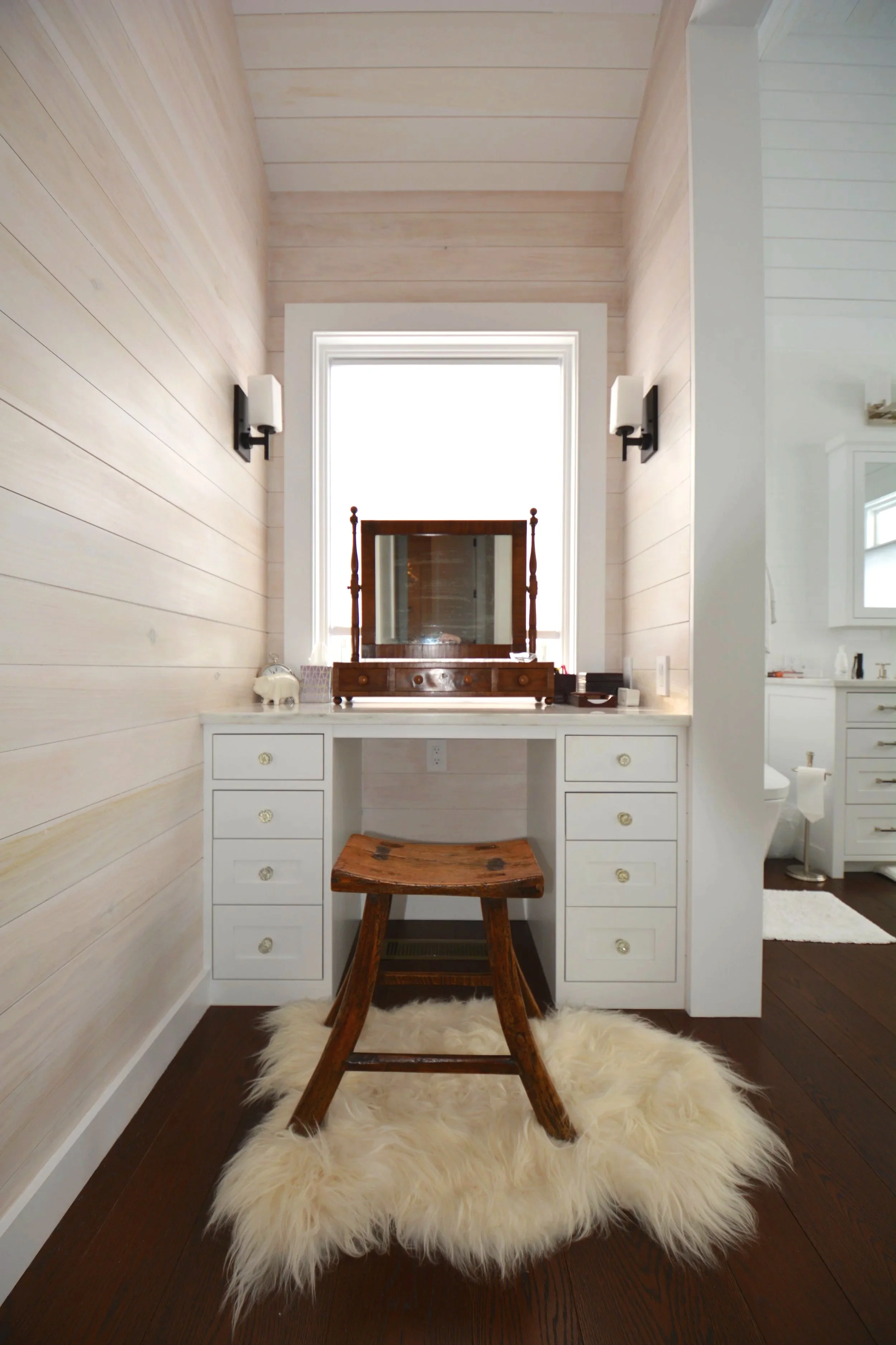 A vanity area with a wooden stool on a fluffy cream-colored rug, a white desk with drawers, a mirror, and a window behind the mirror, with two wall sconces on either side.