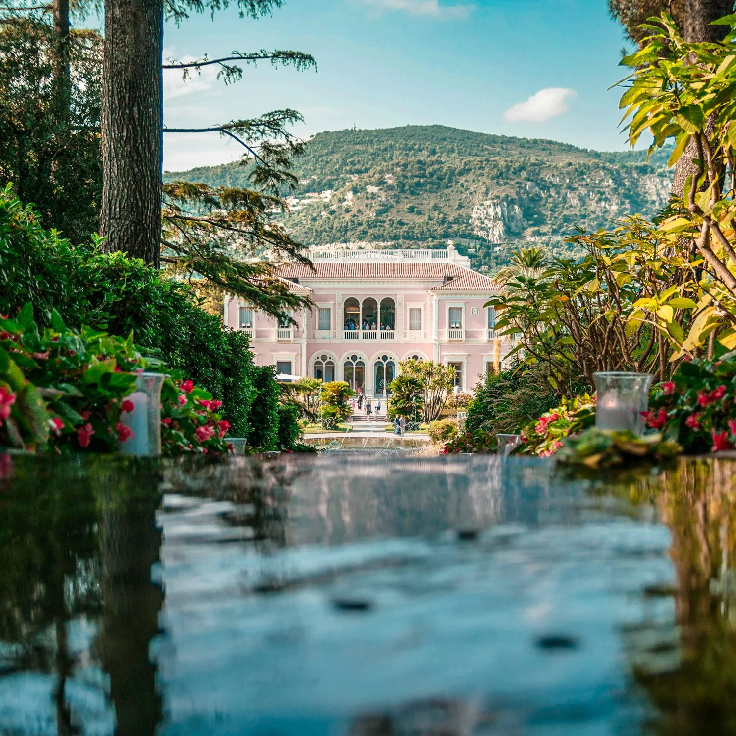 A pink mansion with arched windows and balconies, viewed through a garden with flowers and trees, with mountains in the background and candles on a table in the foreground.