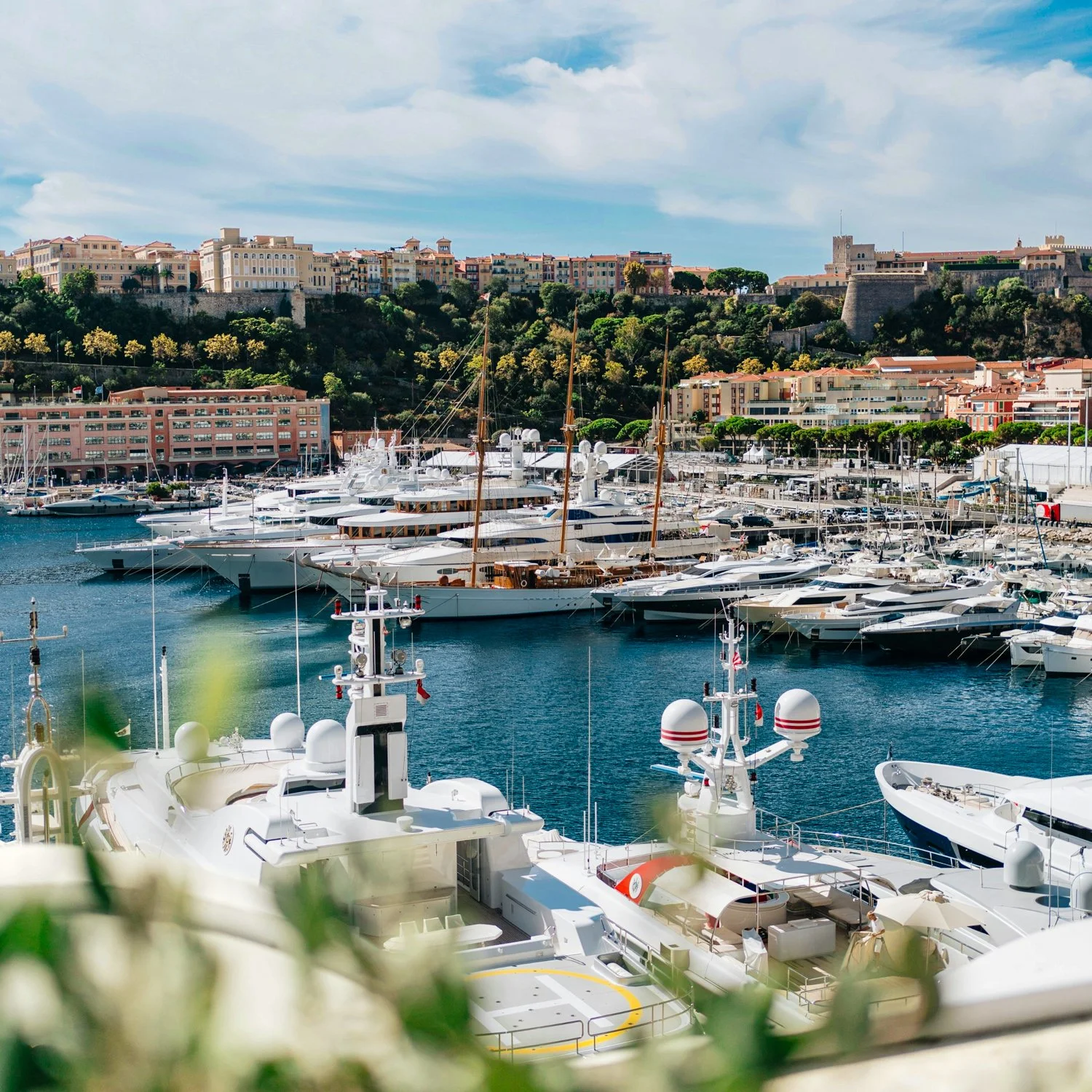 Marina filled with luxury yachts and sailboats, with hillside buildings and greenery in the background, under a partly cloudy sky.